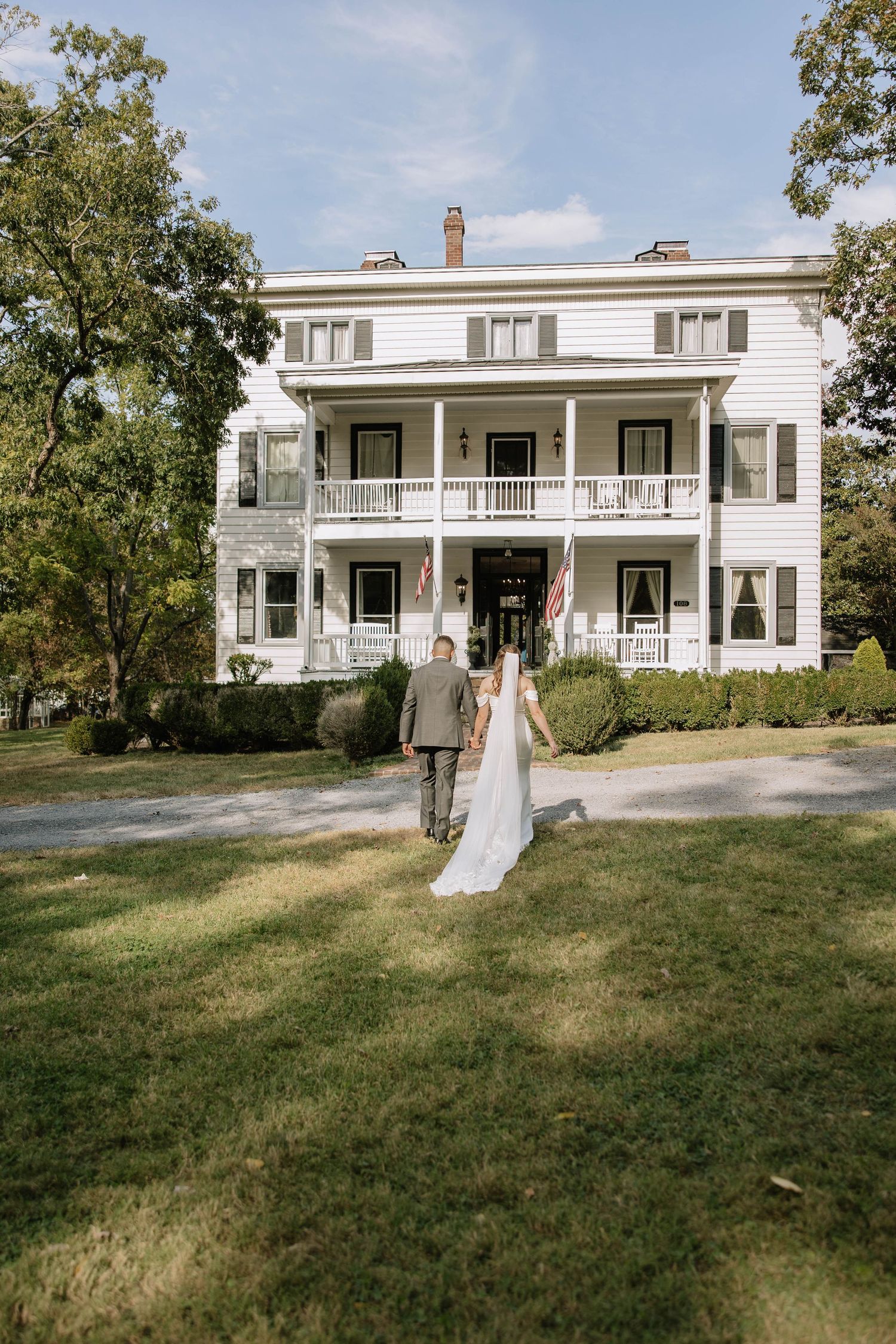 A white historic farmhouse with wraparound porch serves as an elegant backdrop for a rustic outdoor wedding scene.