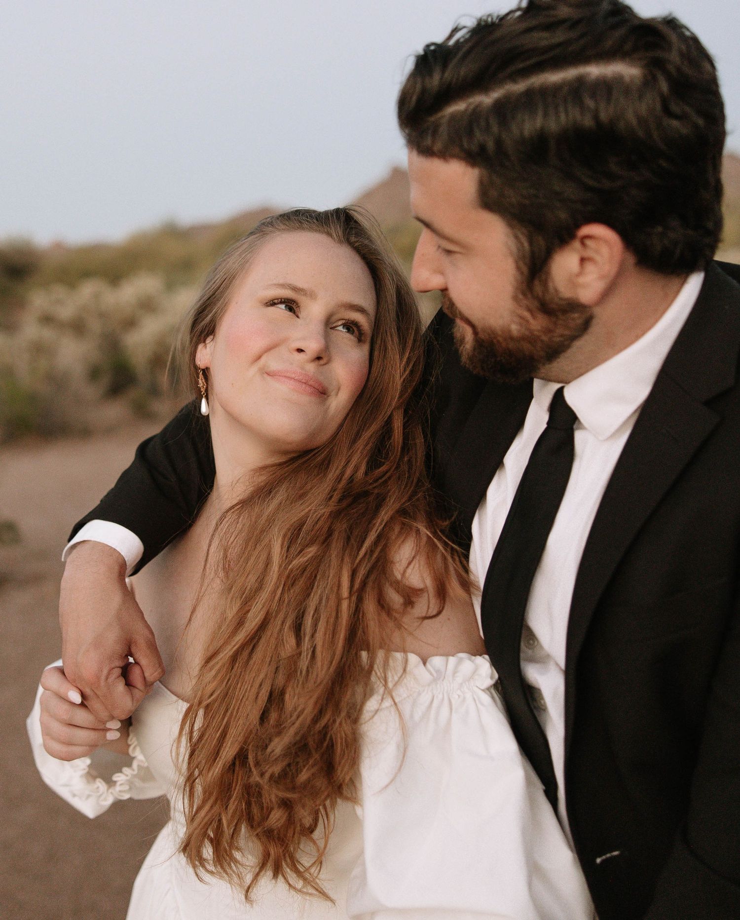 A couple in wedding attire smiles at each other outdoors, the bride in white and groom in a black suit with tie.