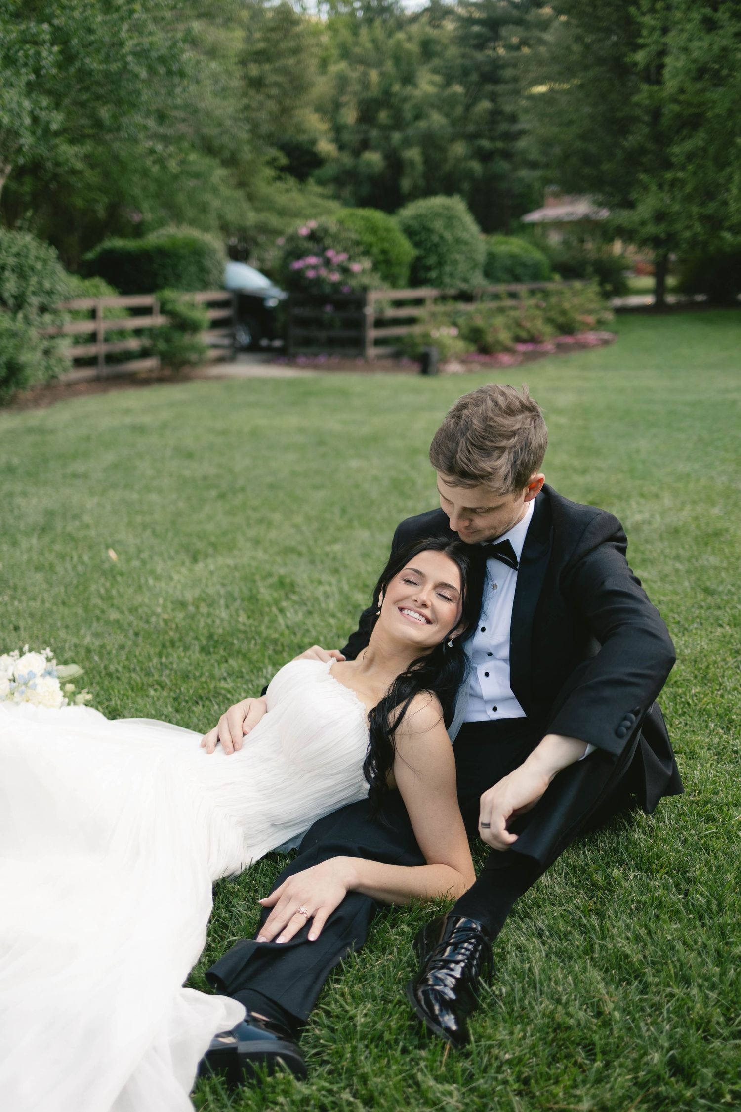 Bride in white gown and groom in black tuxedo sitting together on green grass, smiling, with lush garden backdrop.