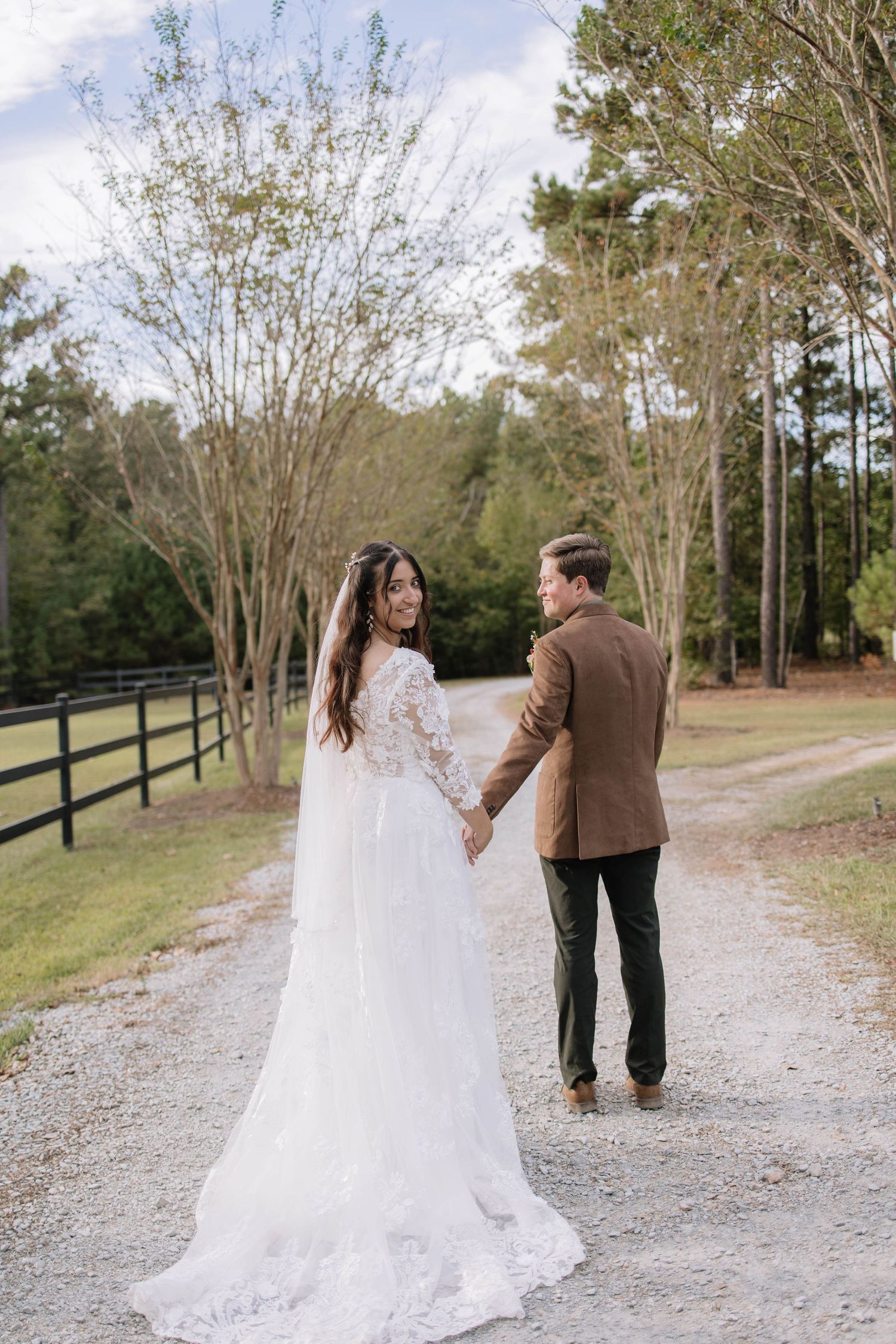 Newlyweds stroll together on a winding gravel path with black fence and trees in background.