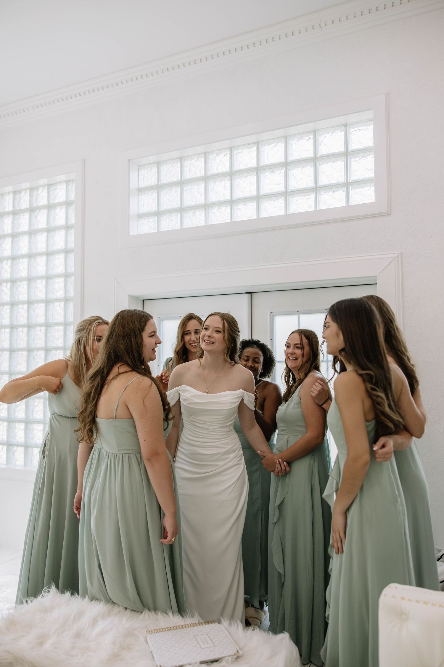 Bridesmaids in sage green dresses gather around the bride in a bright indoor setting.