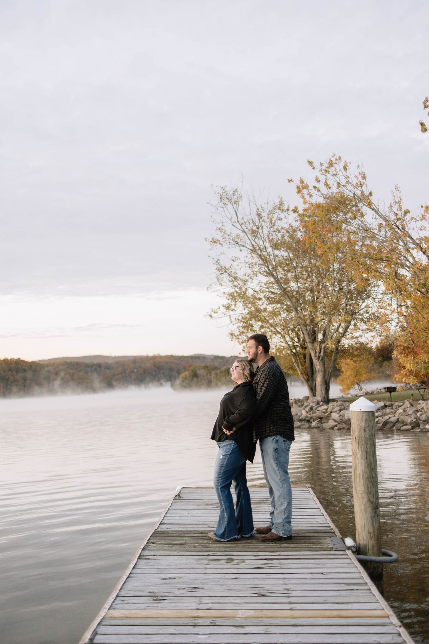 A couple standing on a lakeside dock during autumn with misty water and trees.