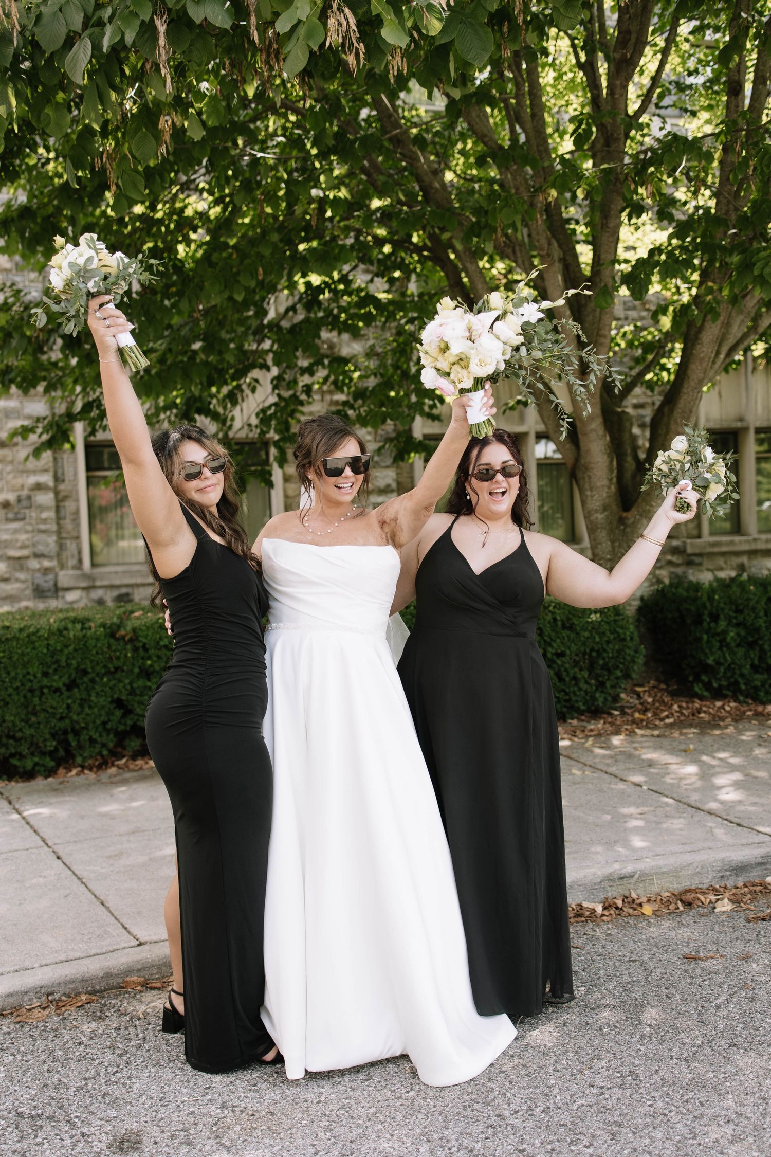 Bridesmaids in black dresses celebrate with bride in white dress on gravel path.
