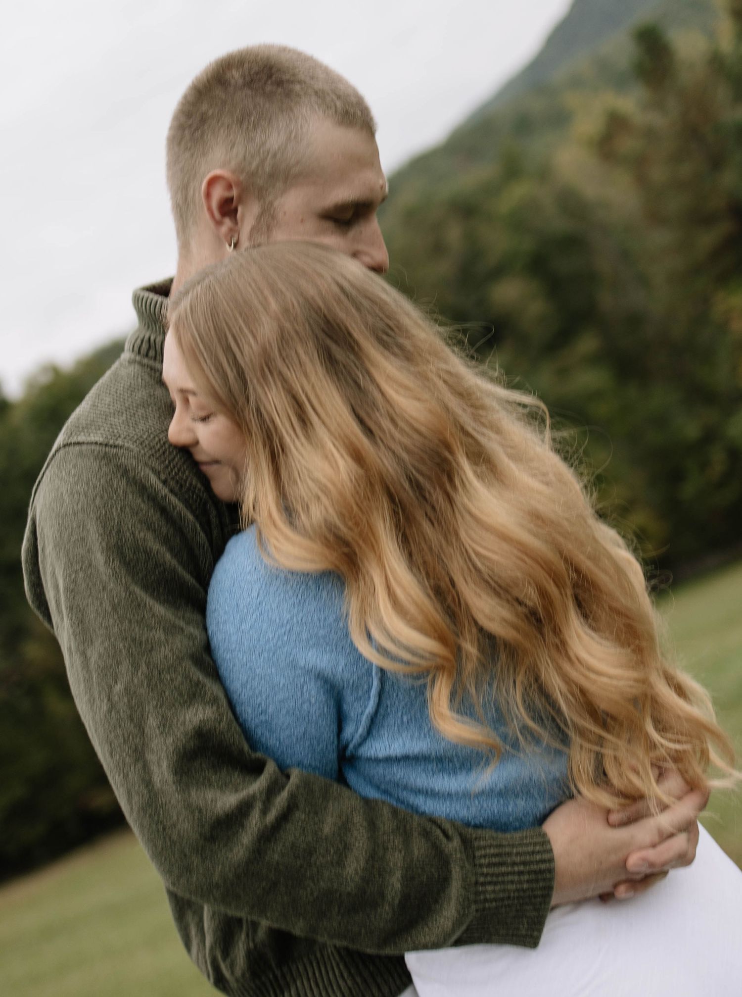 Couple embraces tenderly in a romantic outdoor photoshoot wearing blue and olive green sweaters.