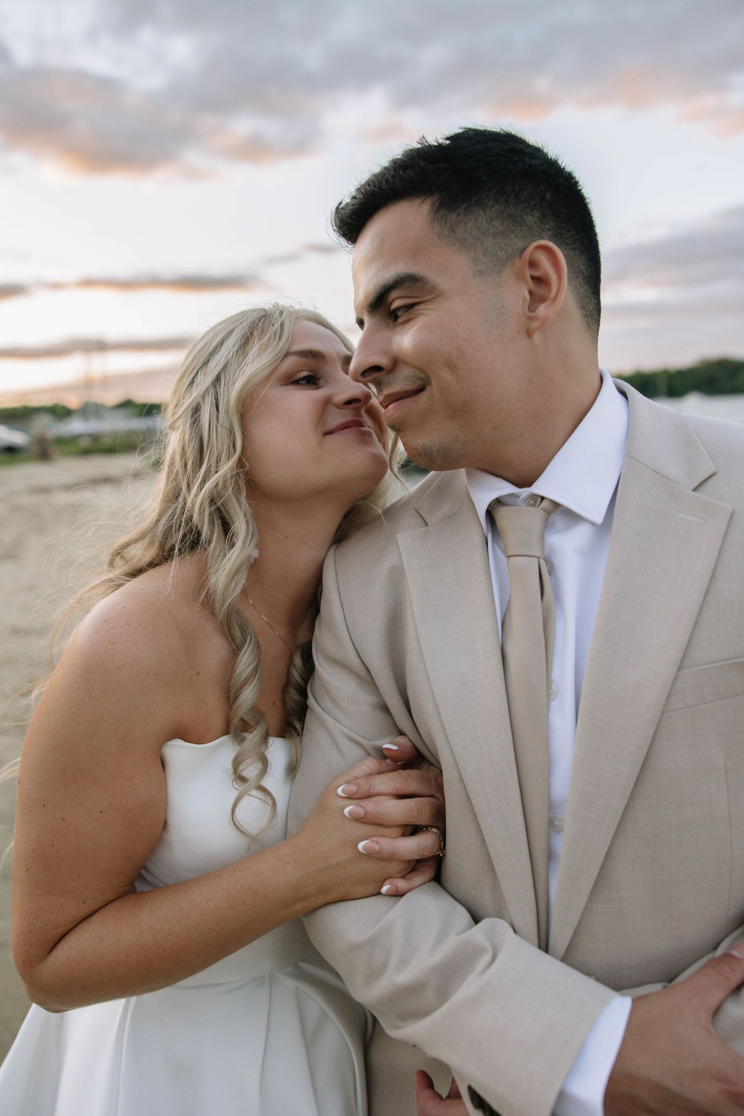 Bride and groom embrace at sunset on a beach, she in white dress, he in beige suit with tie.