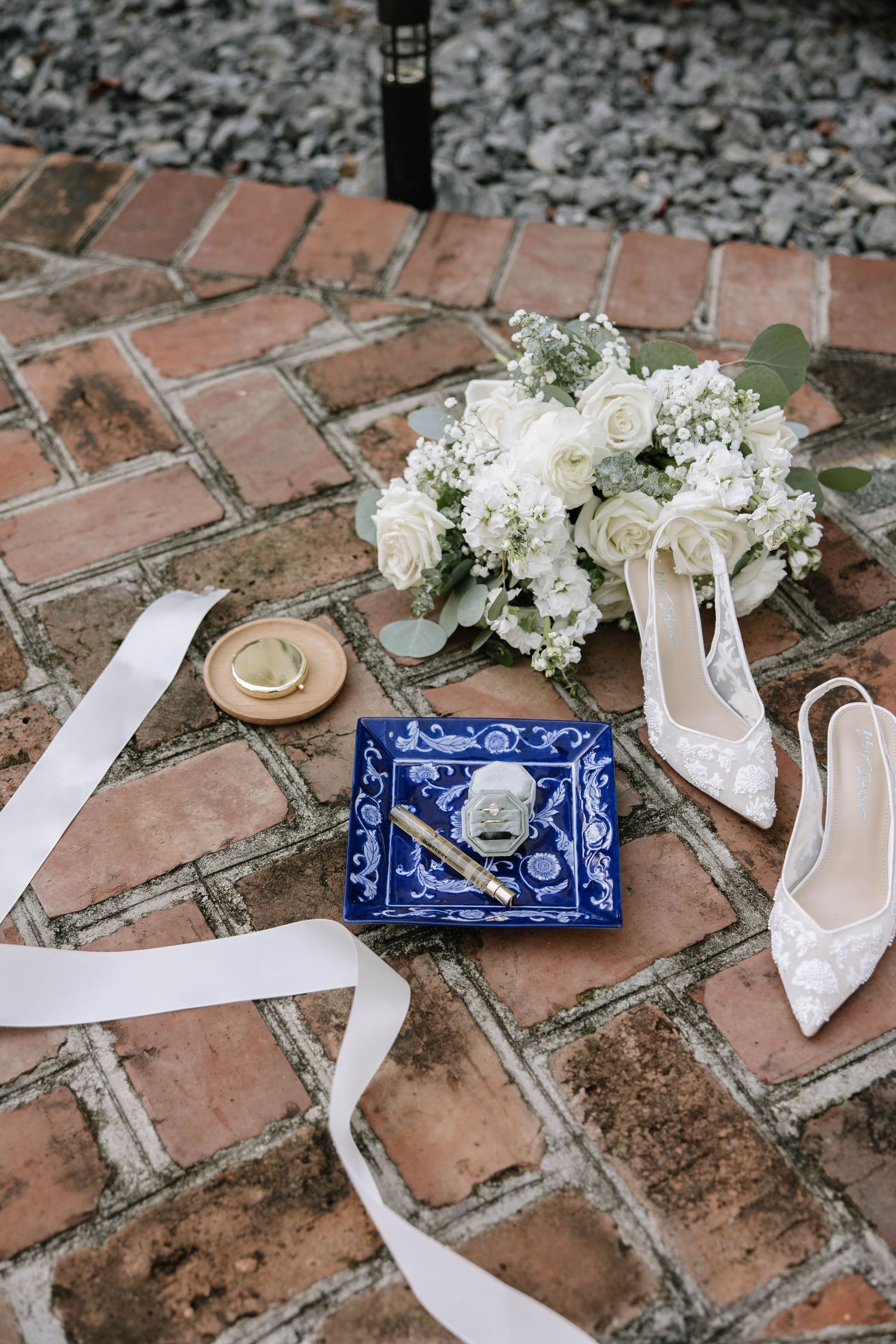 White wedding shoes and floral accessories arranged on rustic brick pavement with a blue decorative plate.