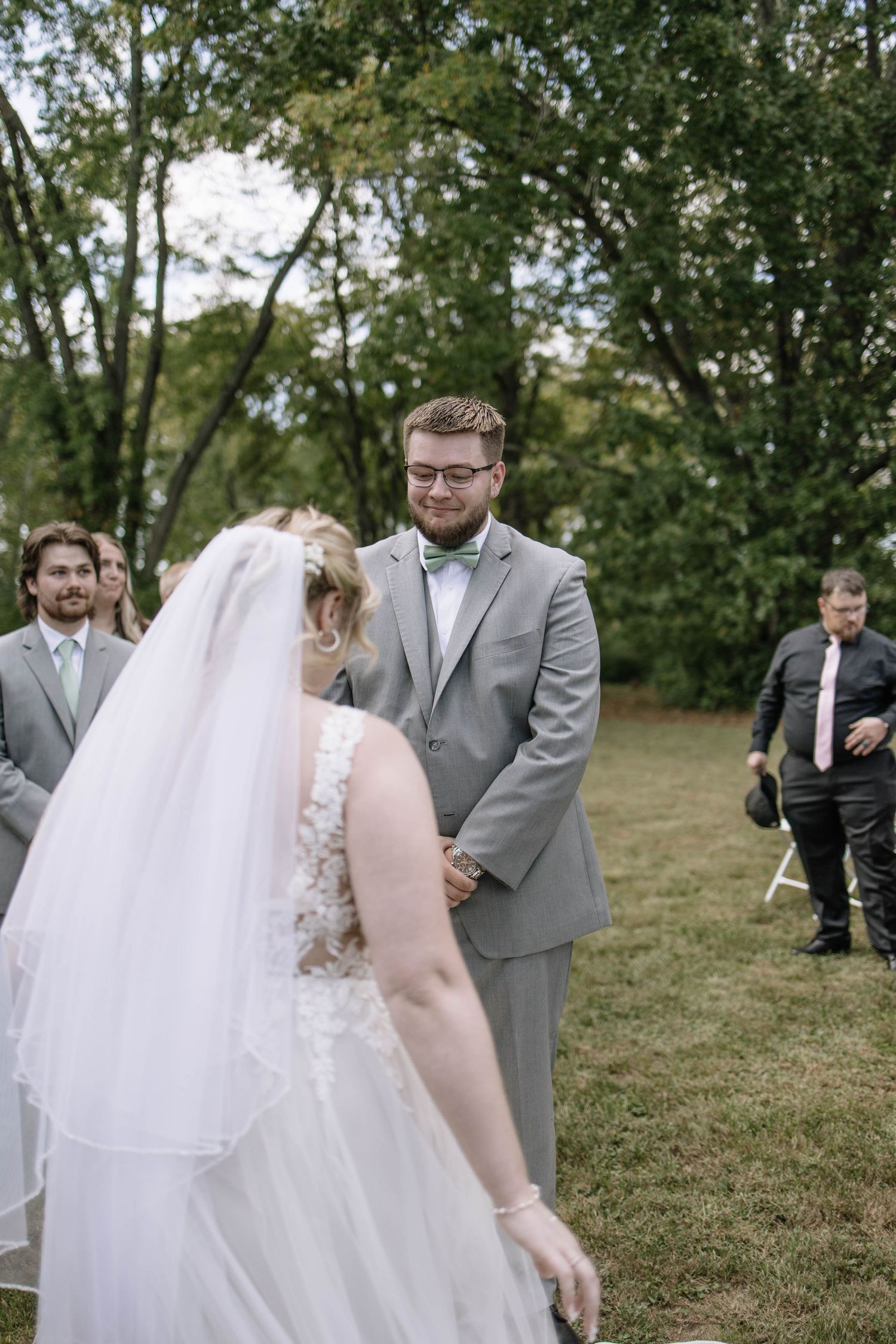Wedding party members react with joy during an outdoor ceremony on a grassy hill.