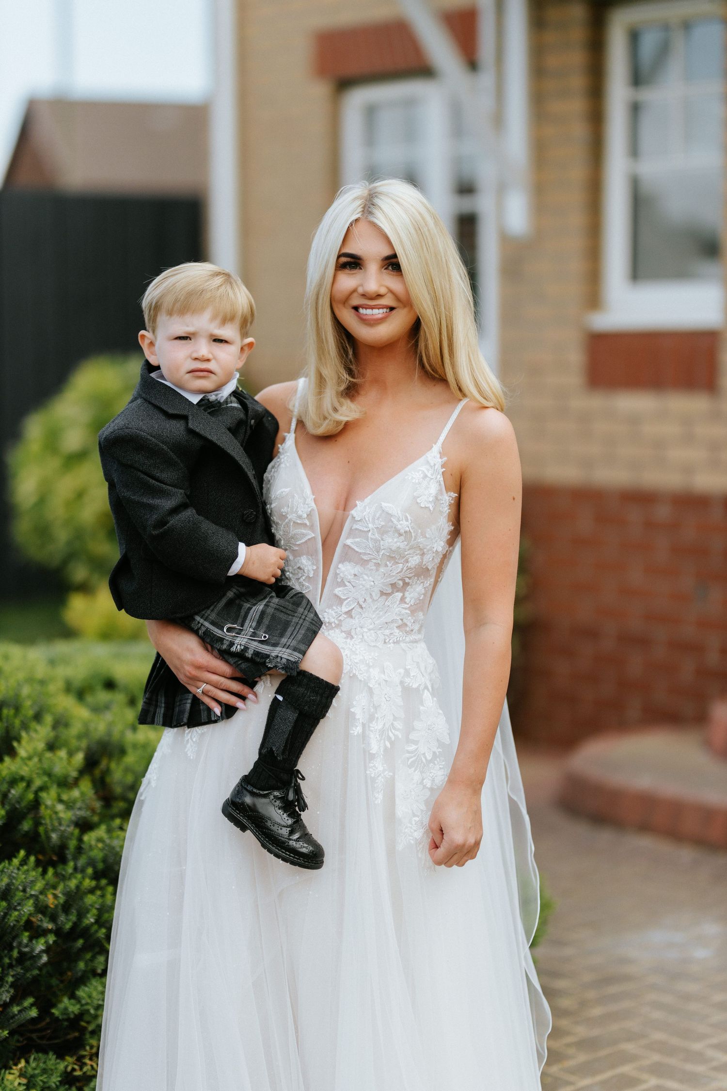 A person in a white lace wedding dress holds a young child wearing a black formal outfit outside a brick building.