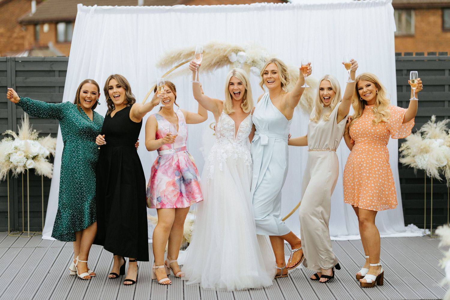 A wedding party celebrates together on a decorated outdoor deck with white floral arrangements in the background.