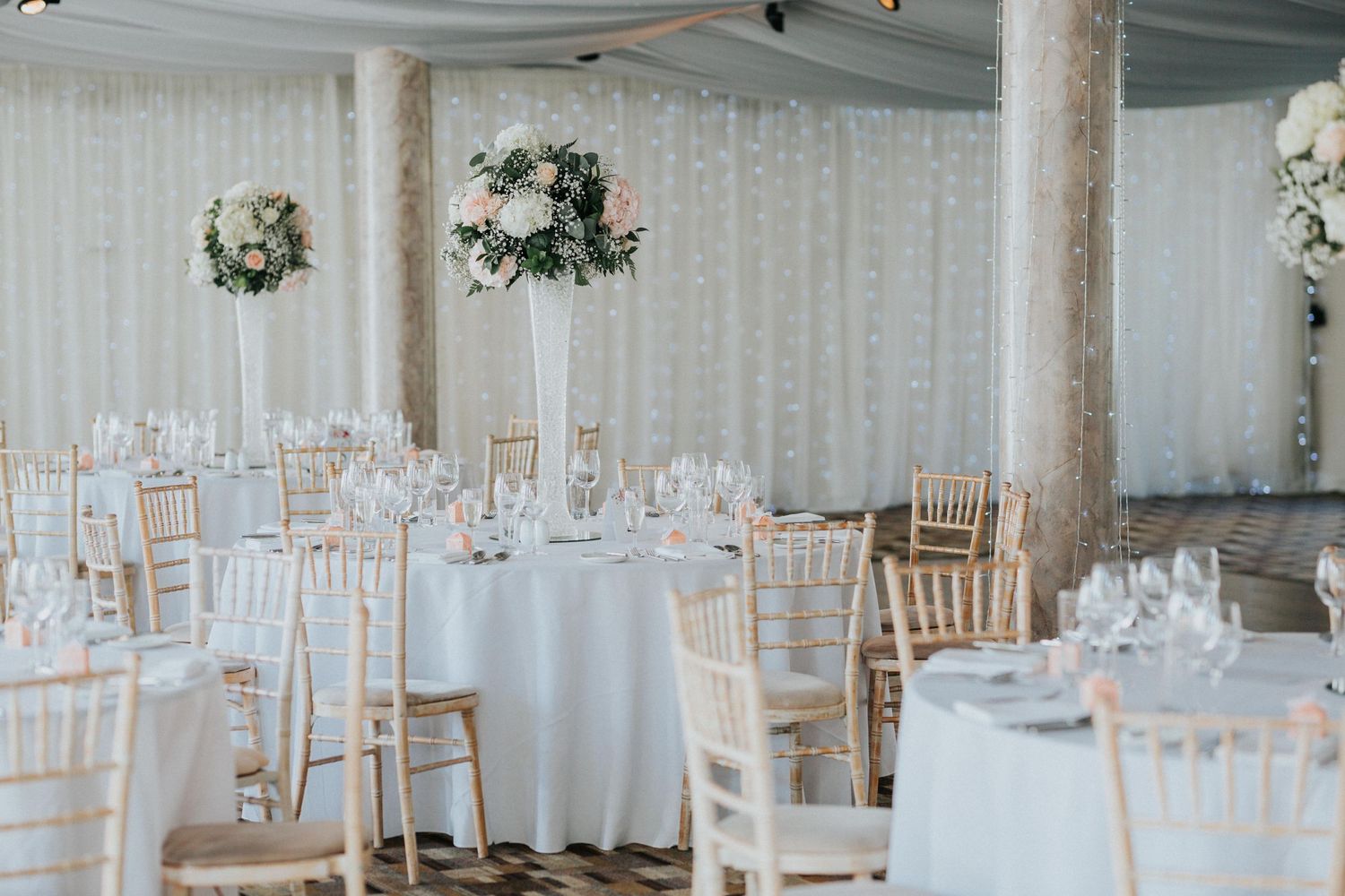 Elegant wedding reception venue with white draped walls, floral wreaths, and chiavari chairs around circular tables.