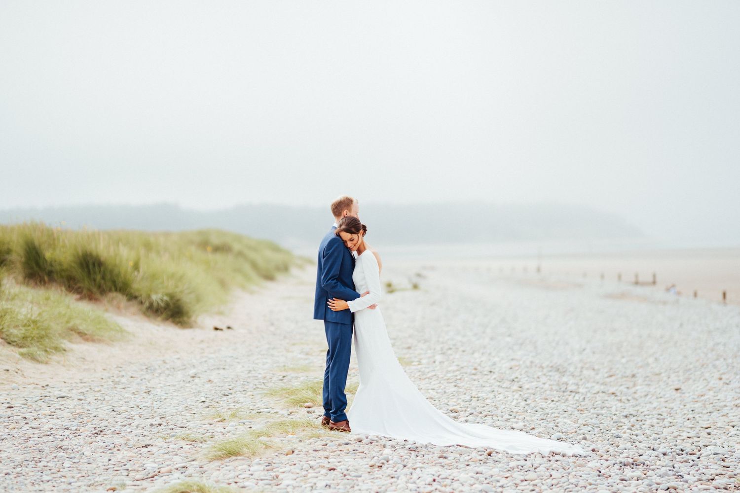 A couple embraces on a misty beach with sand dunes and coastal grass in the background.
