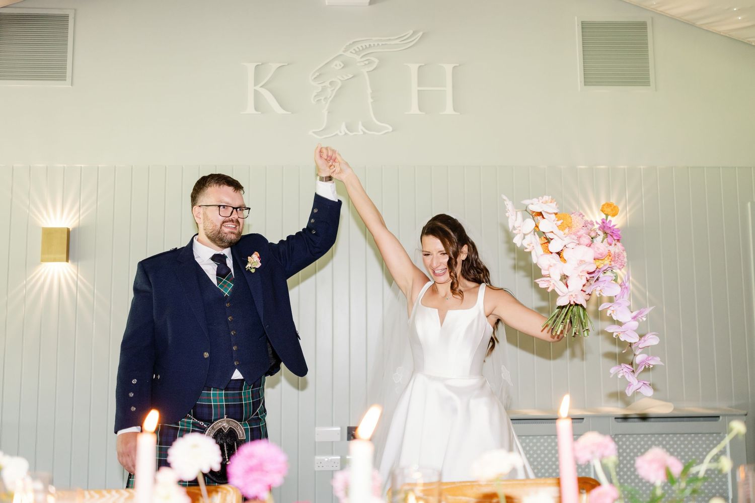 A celebratory moment at a wedding reception with a green backdrop, pink floral decorations and raised arms in triumph.