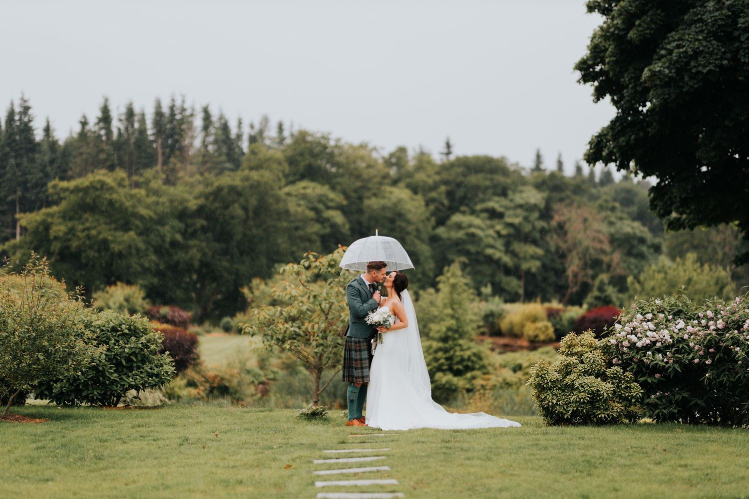 A couple shares a kiss under a white umbrella in a lush garden setting on their wedding day.