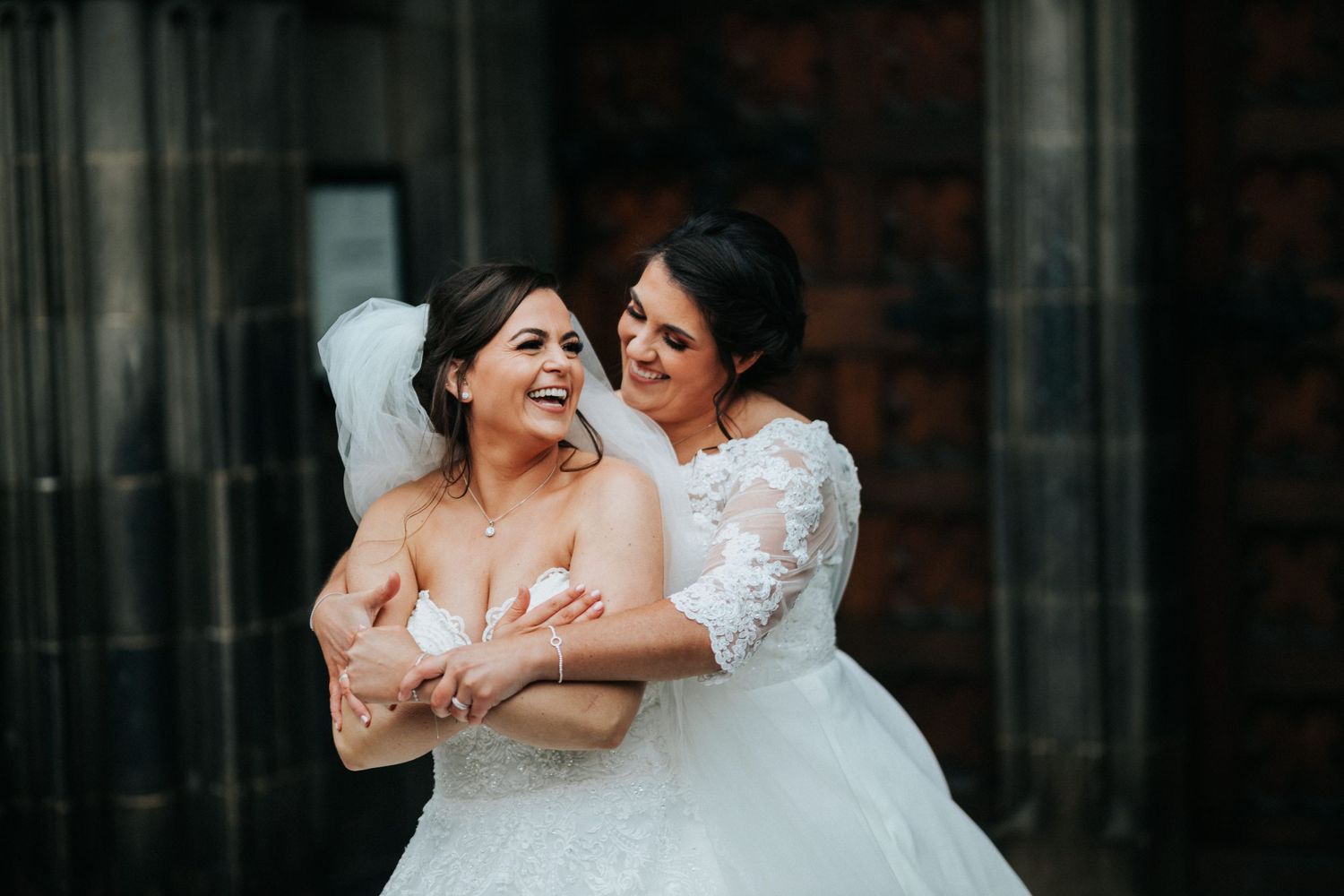 Two brides embrace and laugh together while wearing white wedding dresses against a brick wall backdrop.