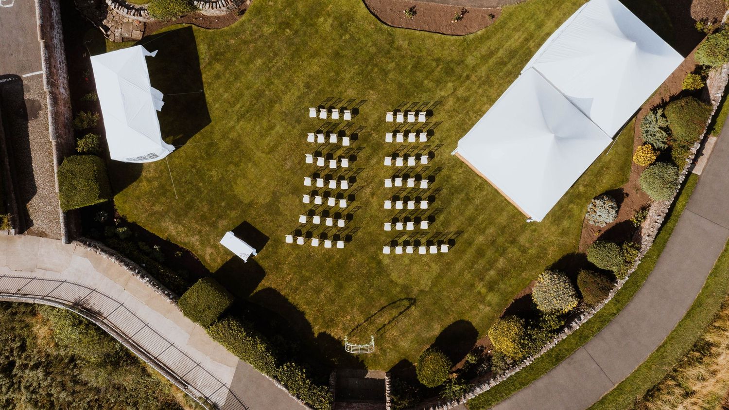 Aerial view of white chairs arranged in rows on a green lawn with event tents and curved pathway visible.