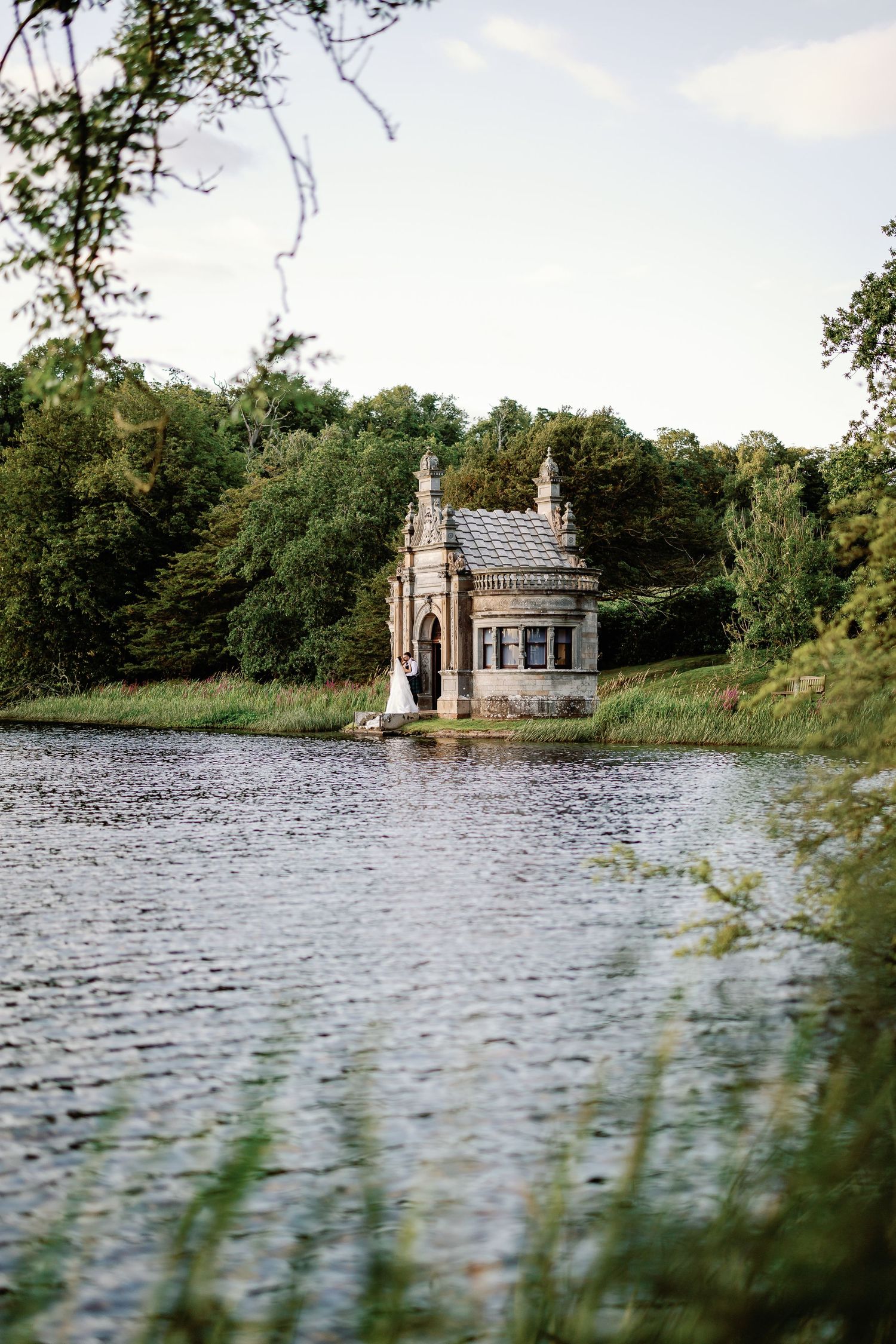 A gothic-style stone boathouse stands partially submerged in a tranquil lake surrounded by lush green trees.