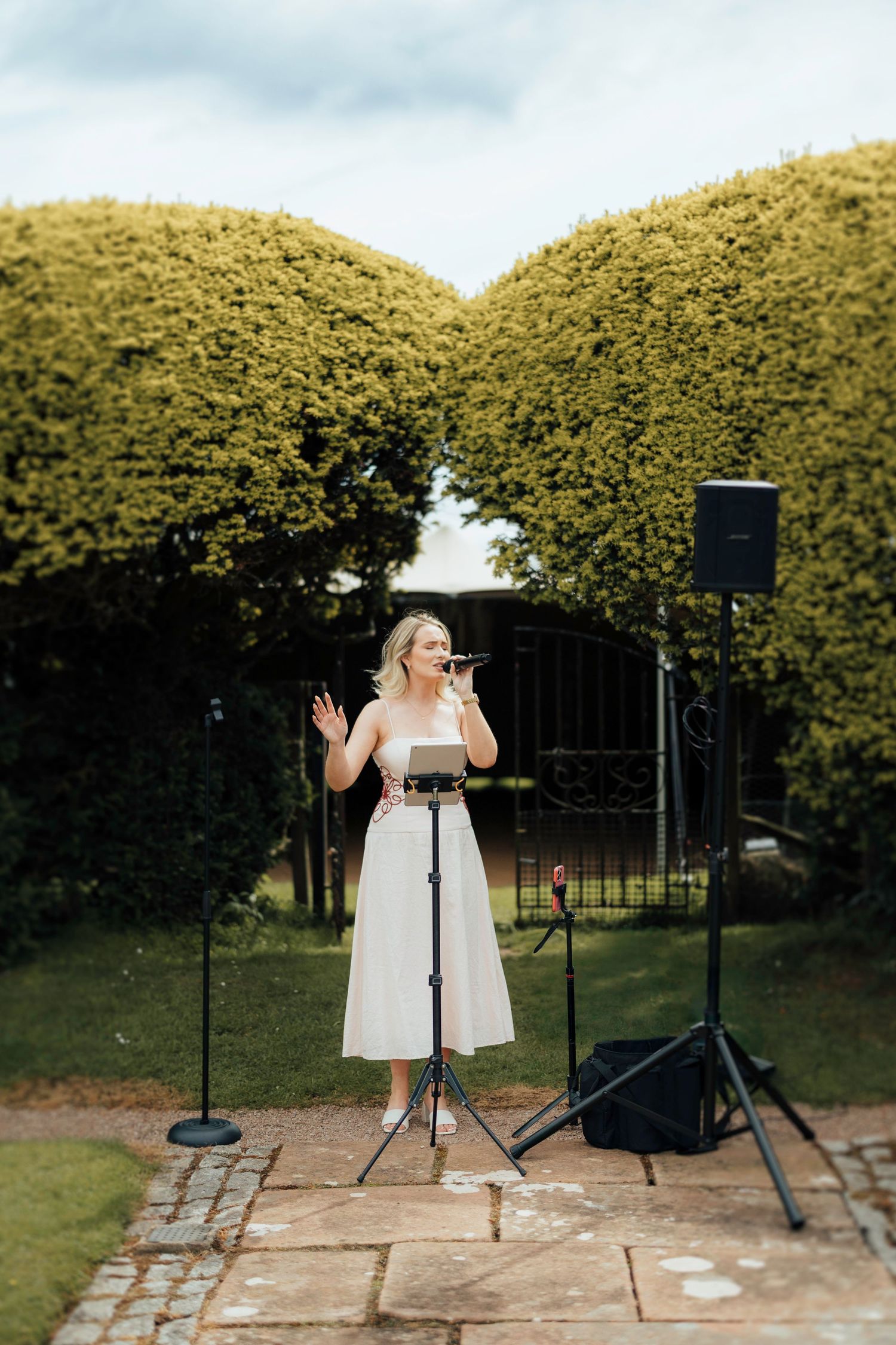 A person in a white dress performs at an outdoor venue with tall manicured hedges in the background.