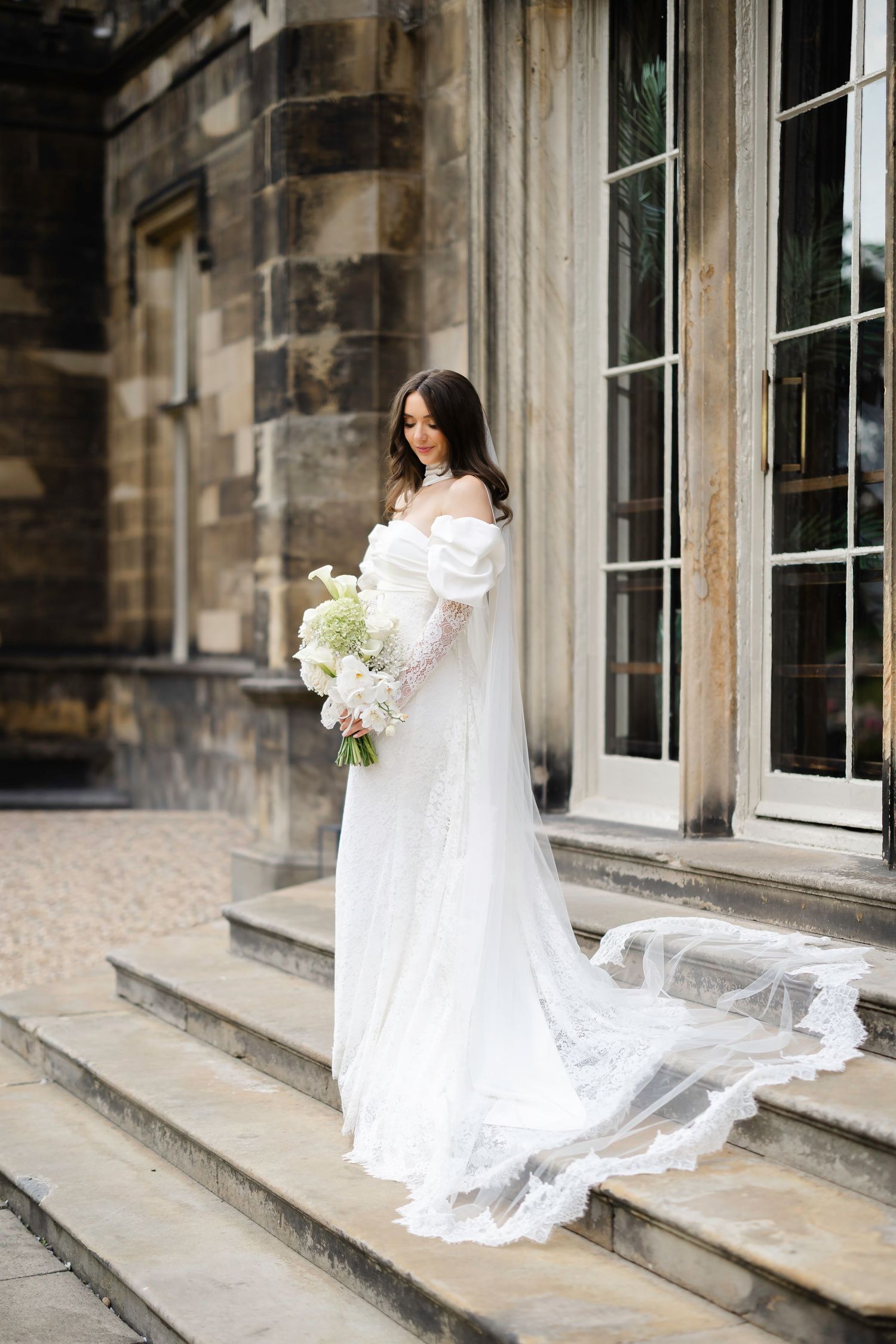 Elegant bride in white off-shoulder gown with lace veil holding bouquet on stone steps of historic venue.