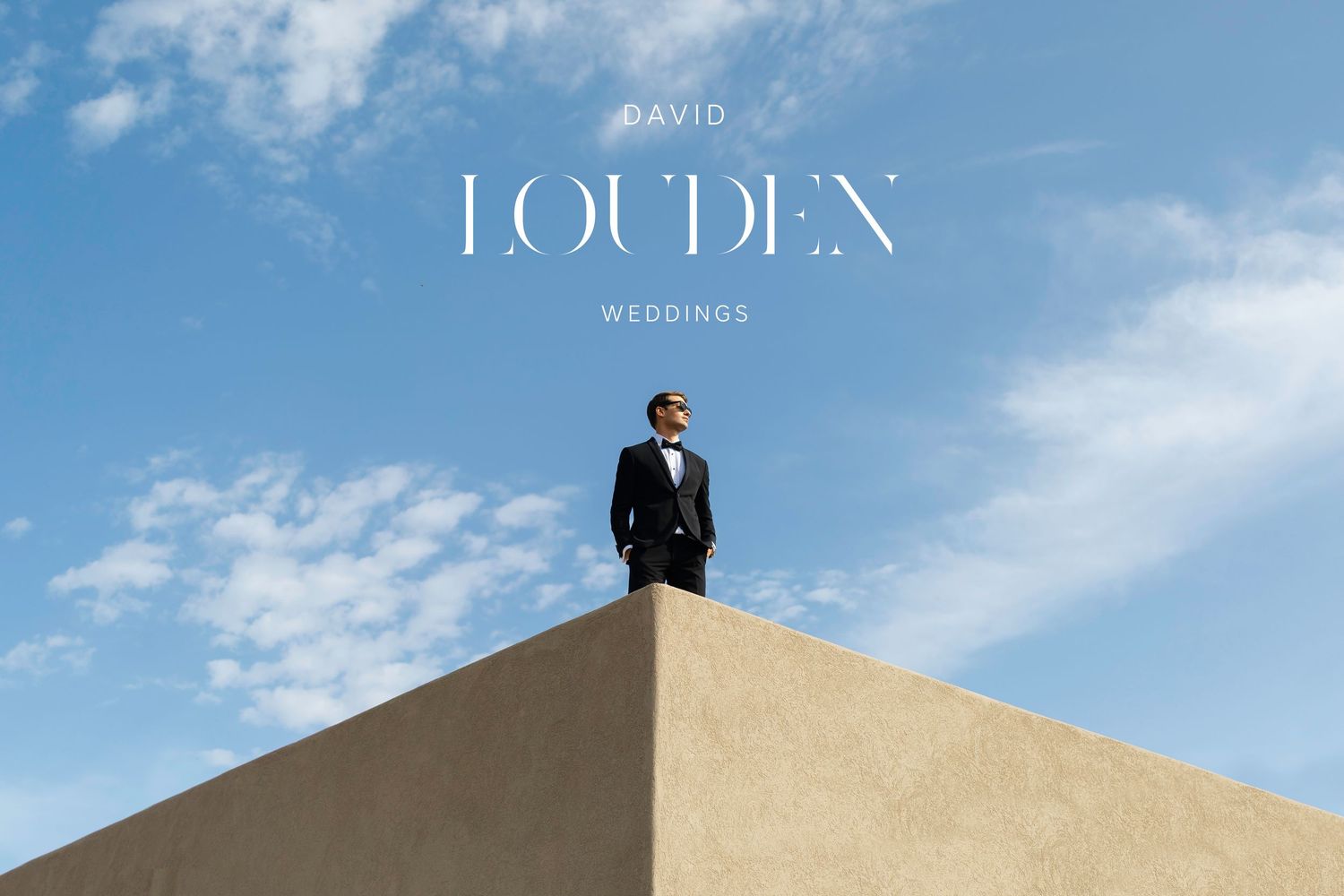A groom in a black tuxedo stands atop a white wall against a blue sky, with David Louden Weddings text overlay.