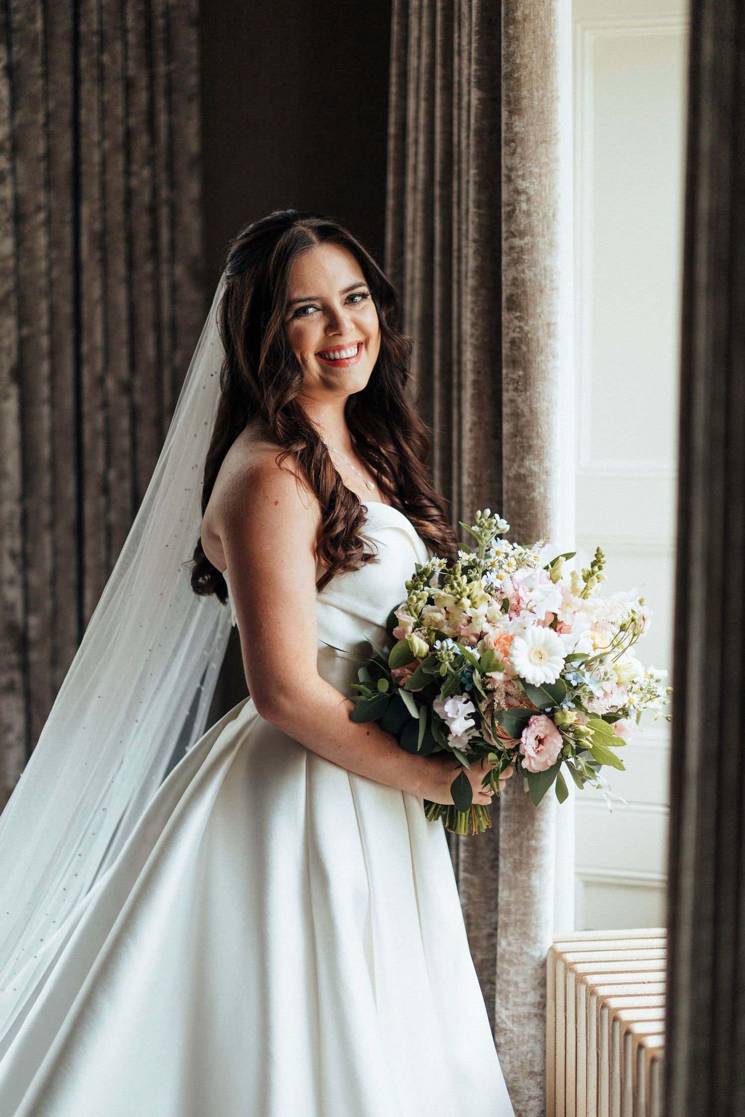 A bride in an elegant white ballgown and long veil holds a beautiful blush and ivory bouquet by a window.