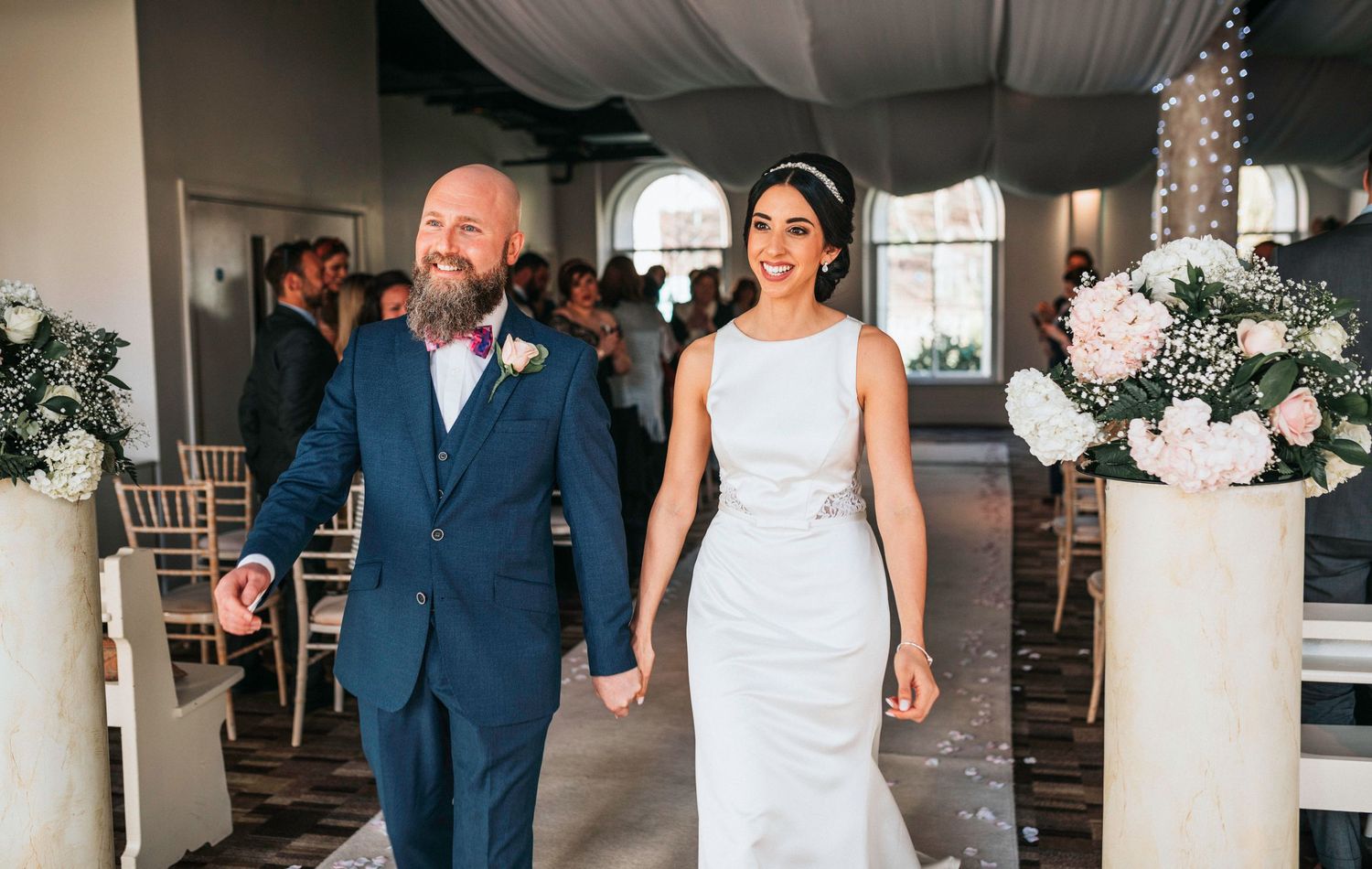 Happy couple walks hand in hand at their elegant indoor wedding ceremony with white floral decor and chiavari chairs.