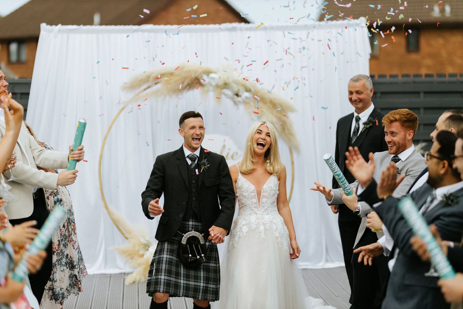 Wedding couple walks down aisle under floral arch as guests throw confetti during outdoor Scottish ceremony.