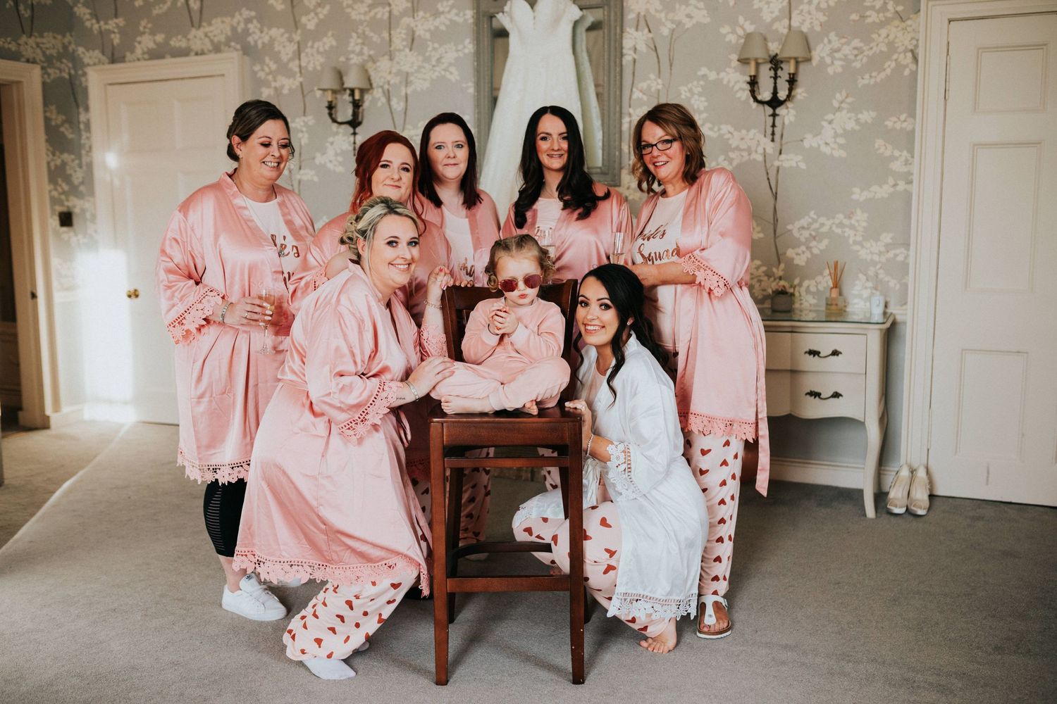 A wedding party in matching pink satin robes poses together in an elegant room with white walls and classic decor.