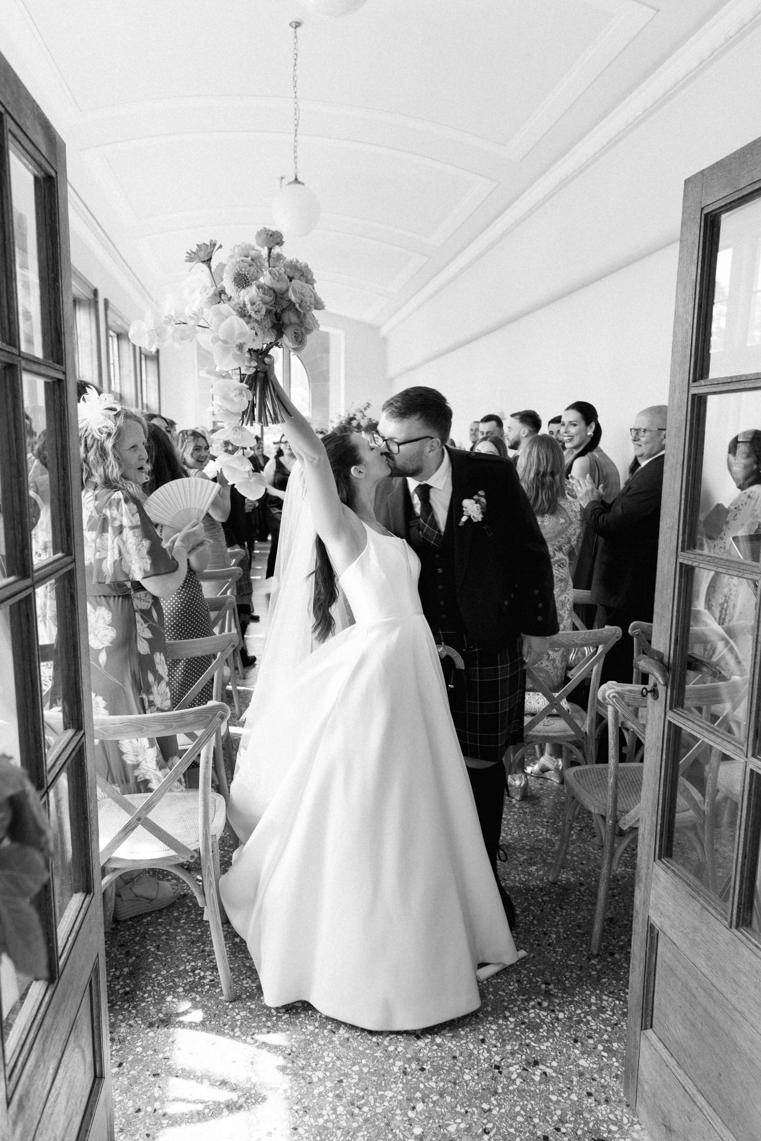A wedding party enters through glass doors into a venue while guests look on and throw confetti during a celebration.