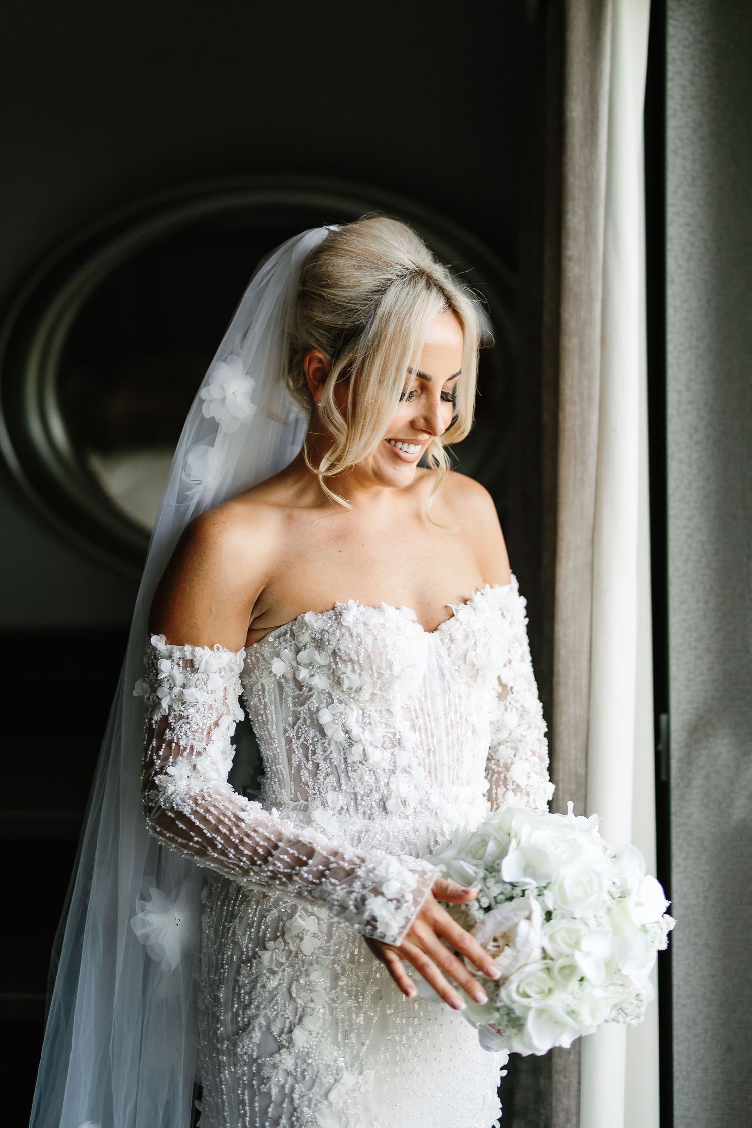 A bride in a delicate lace long-sleeve wedding gown holds a white bouquet while standing by a window.