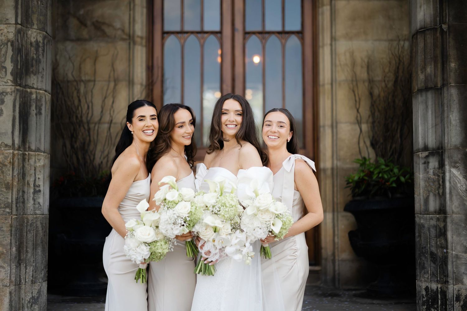 Bride and three bridesmaids in white dresses holding elegant floral bouquets outside a stone church.