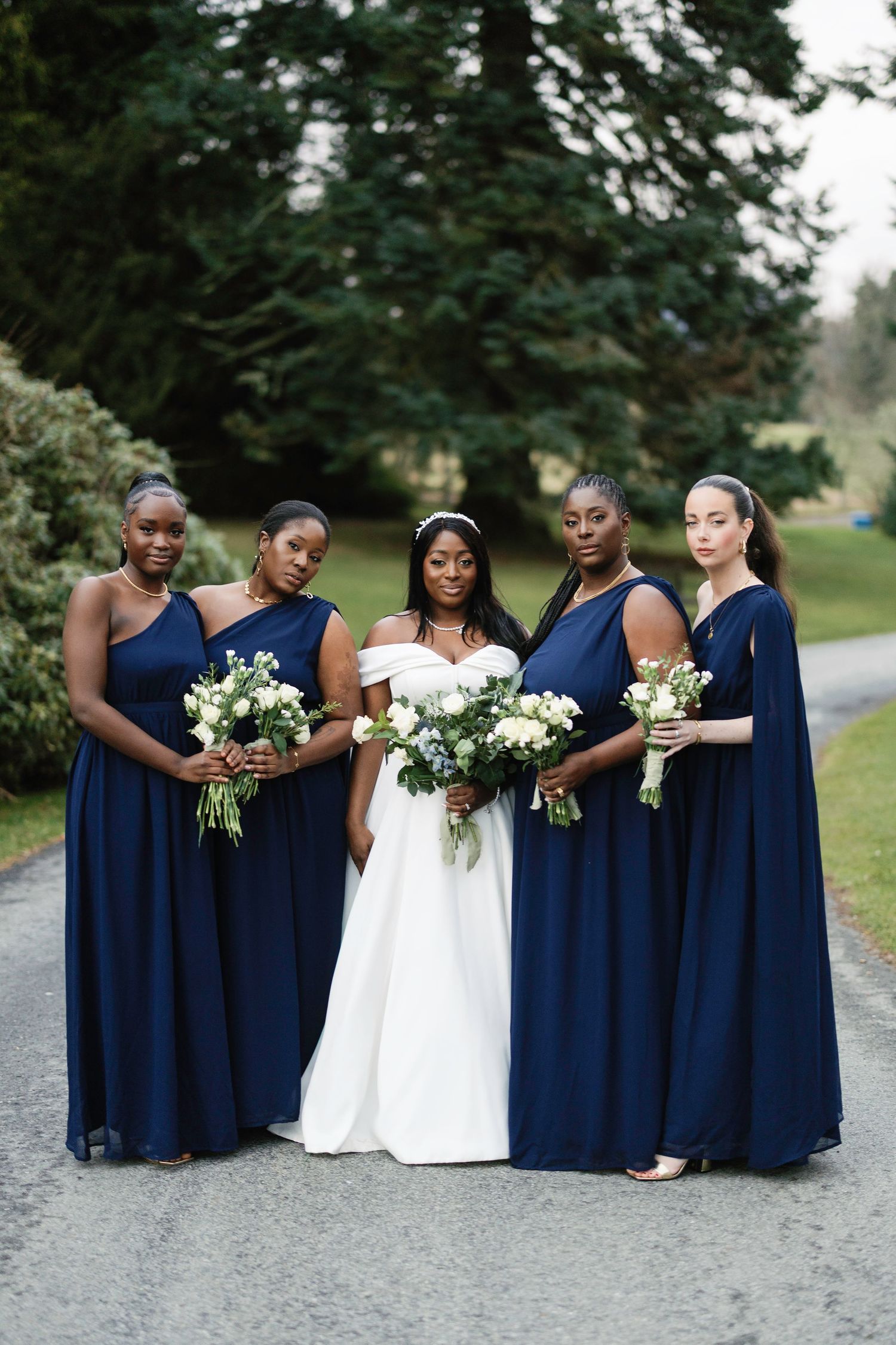 A bridal party wearing elegant navy blue bridesmaid dresses and a white wedding gown pose on a gravel path surrounded by trees.