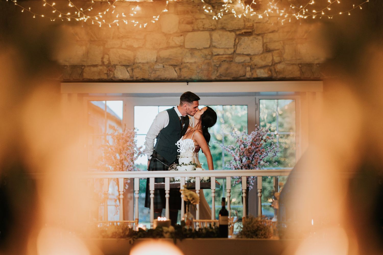 A romantic couple shares an intimate moment on a porch decorated with string lights and floral arrangements against a window backdrop.
