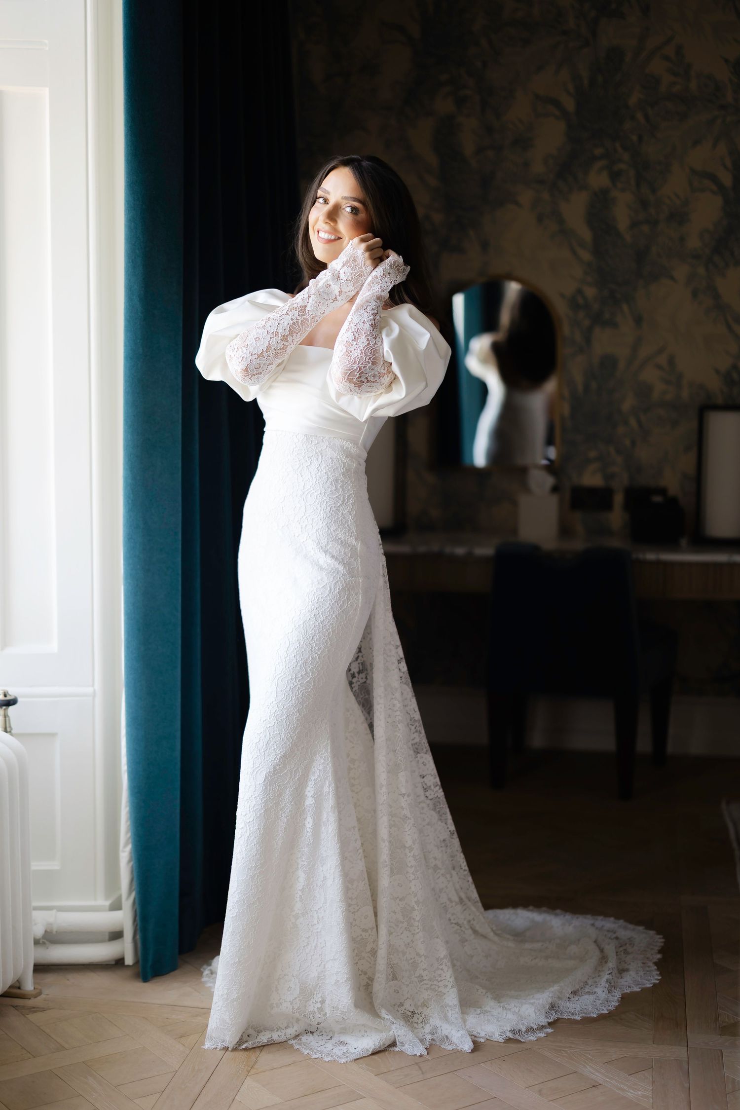 Elegant bride in white lace mermaid wedding dress with puff sleeves and gloves posing in a luxury room.