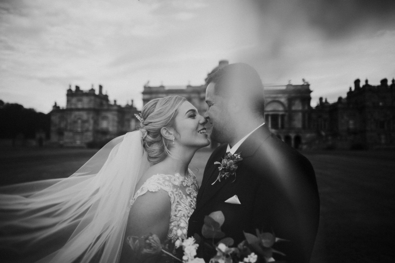 A bride in a flowing white veil and dress embraces their partner in a romantic moment at a stately manor house.