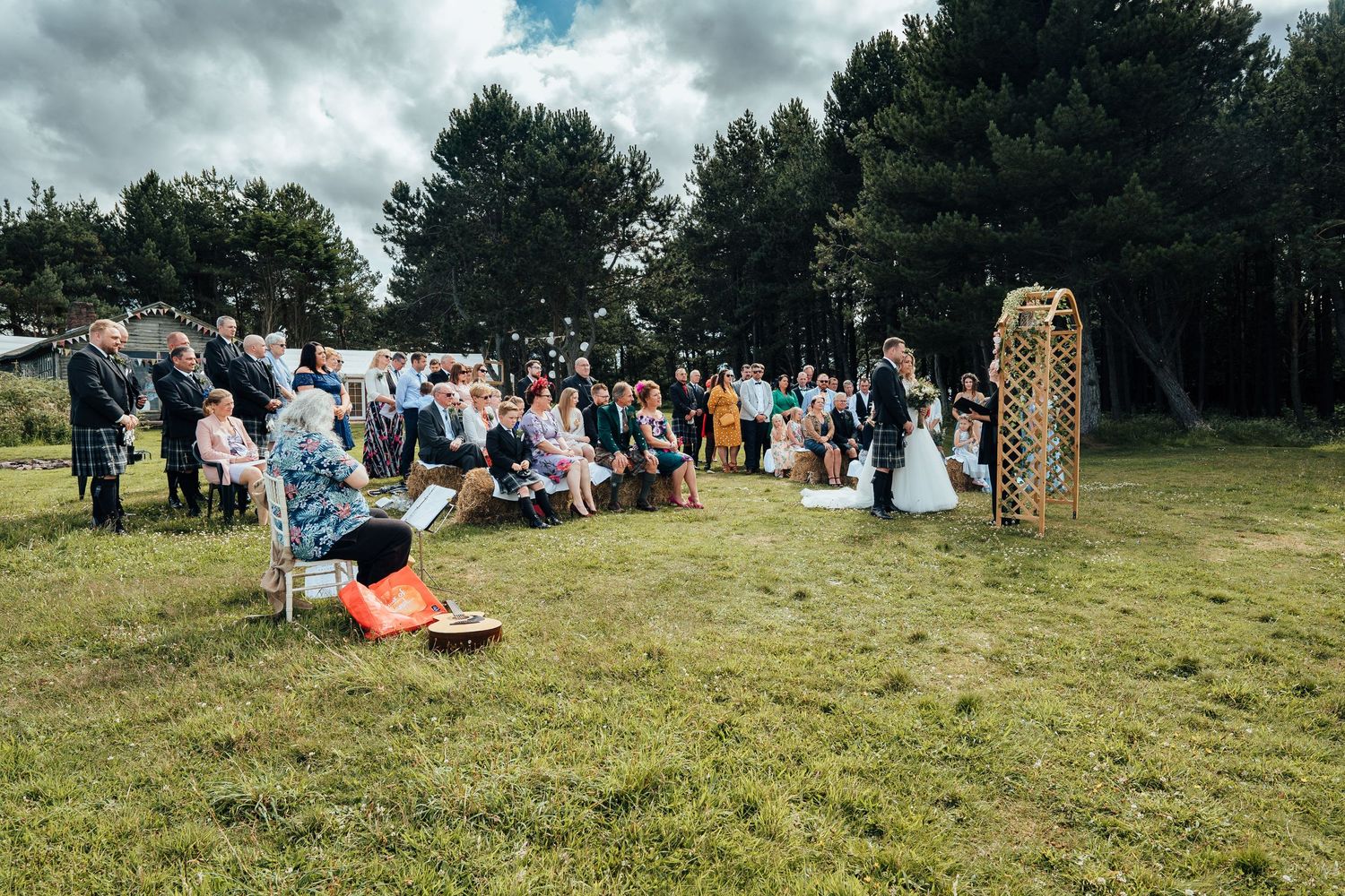 An outdoor wedding ceremony takes place on a grassy field under moody clouds with guests seated and decorative arch.