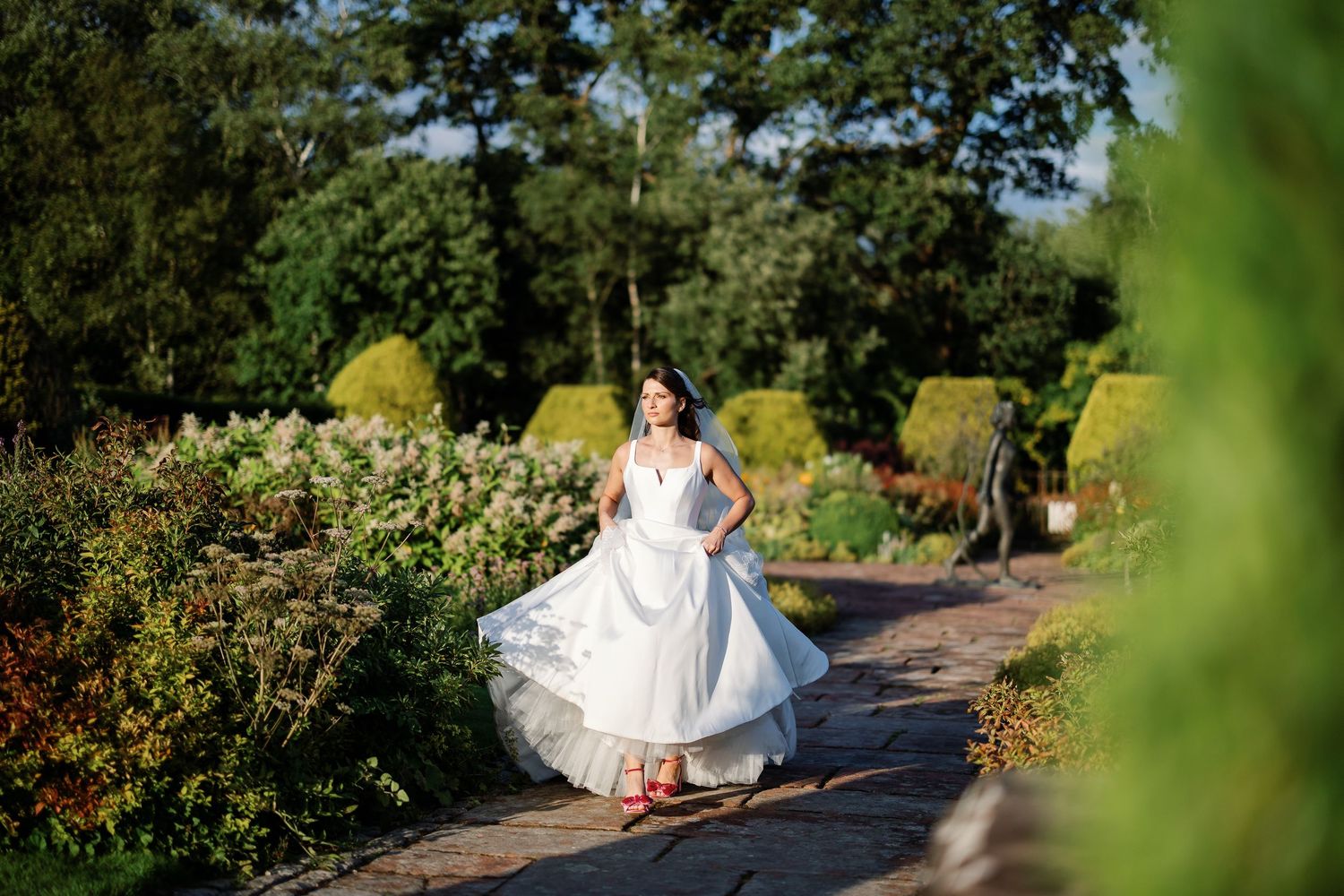 A bride in a flowing white wedding dress walks along a garden path surrounded by lush greenery on a sunny day.