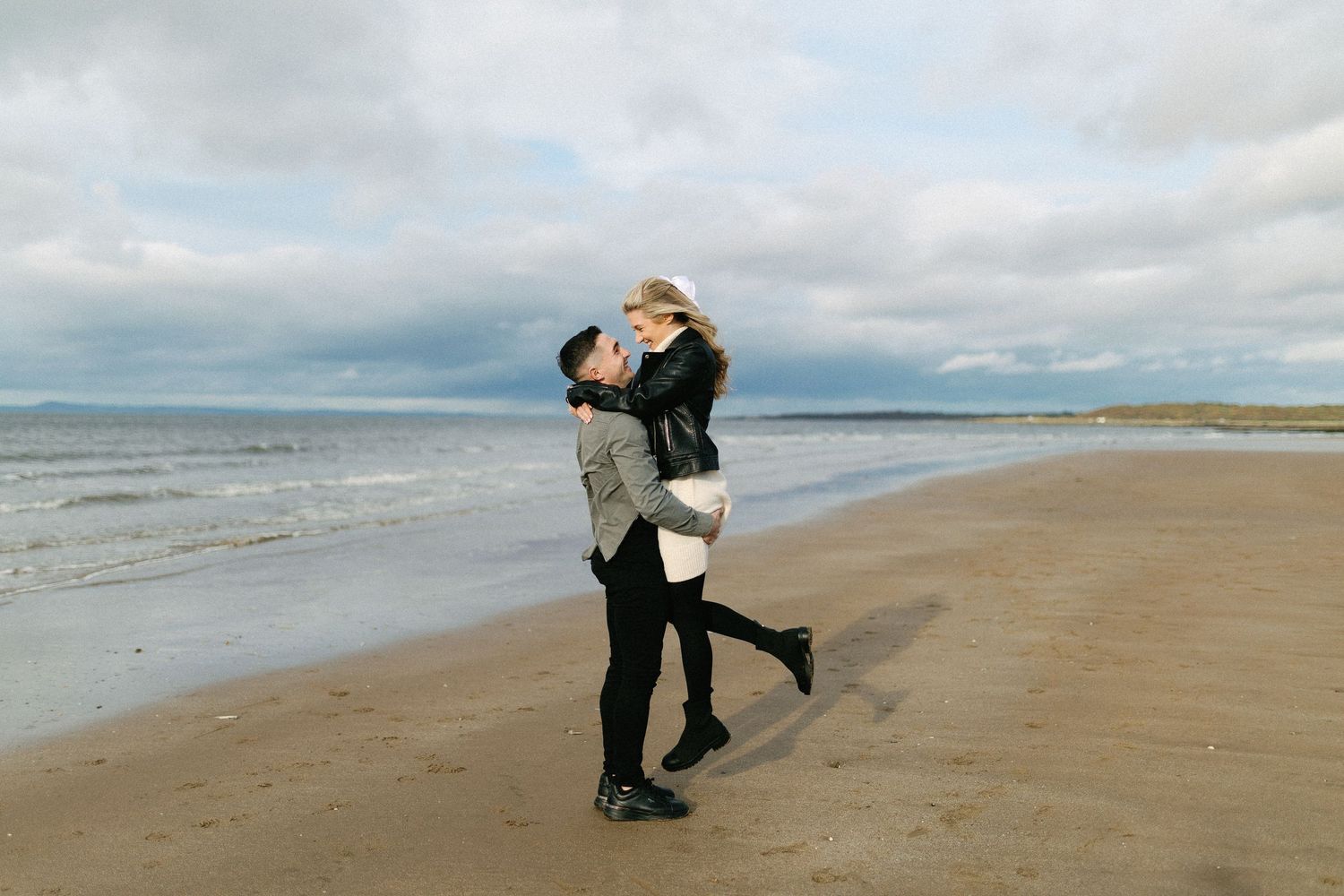 A couple embraces and lifts each other on a cloudy beach with the ocean and grey skies in the background.