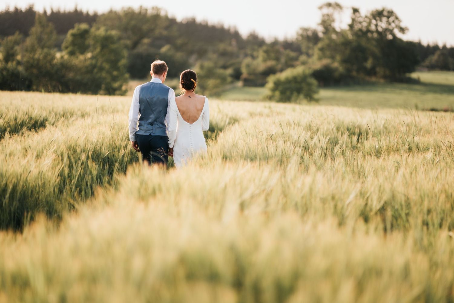 Couple sits together in a sunlit golden wheat field with forest in the background during sunset.