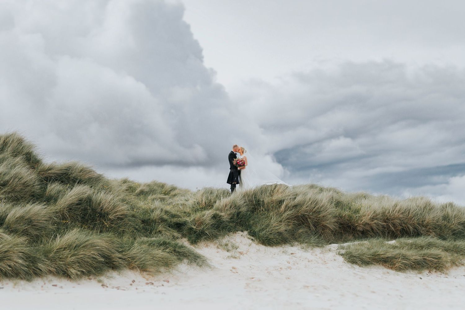 A silhouette stands atop a windswept sand dune with tall grass against a moody cloudy sky.