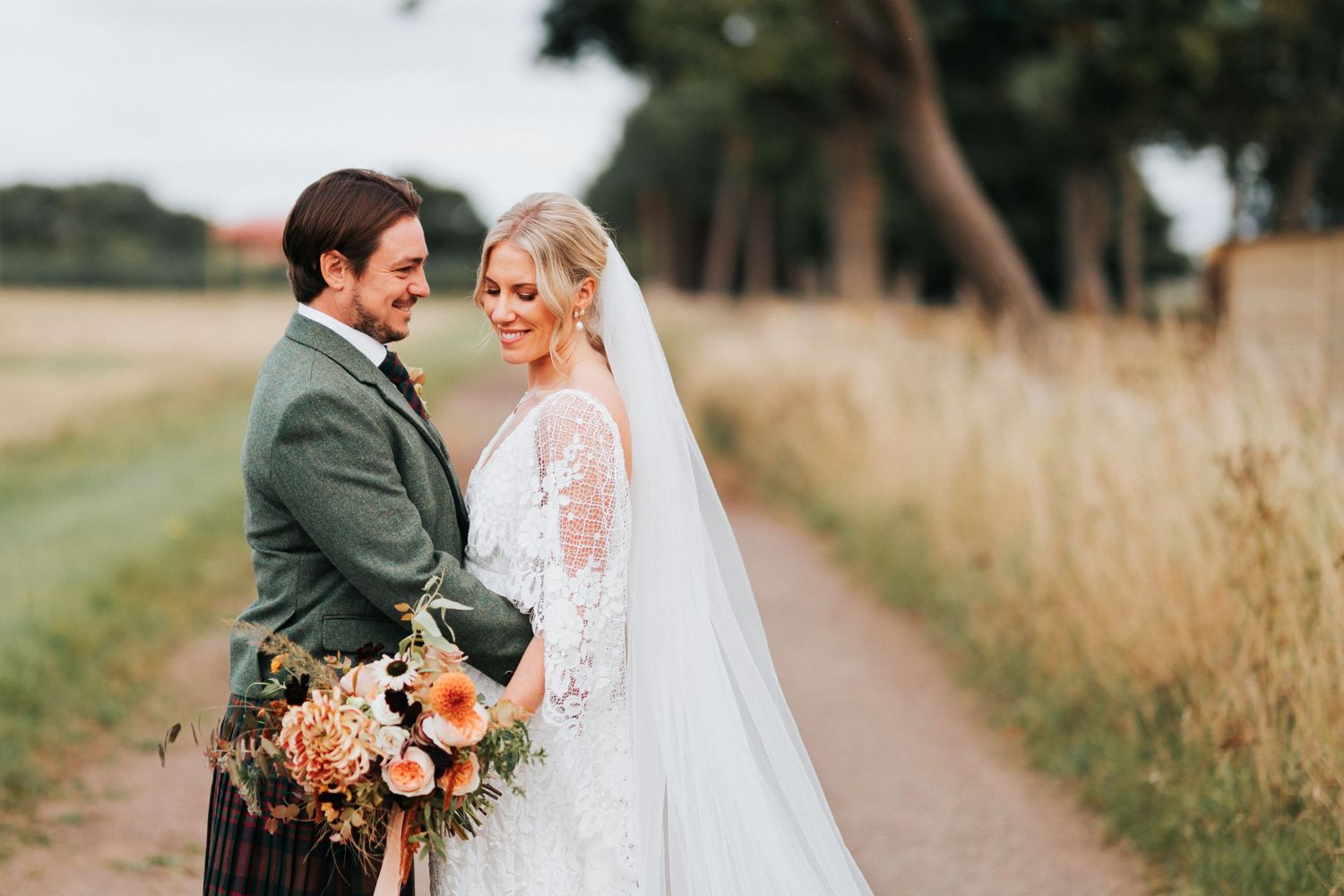 Two happy newlyweds embrace on a country path with the groom wearing a kilt and the bride in a lace wedding dress.