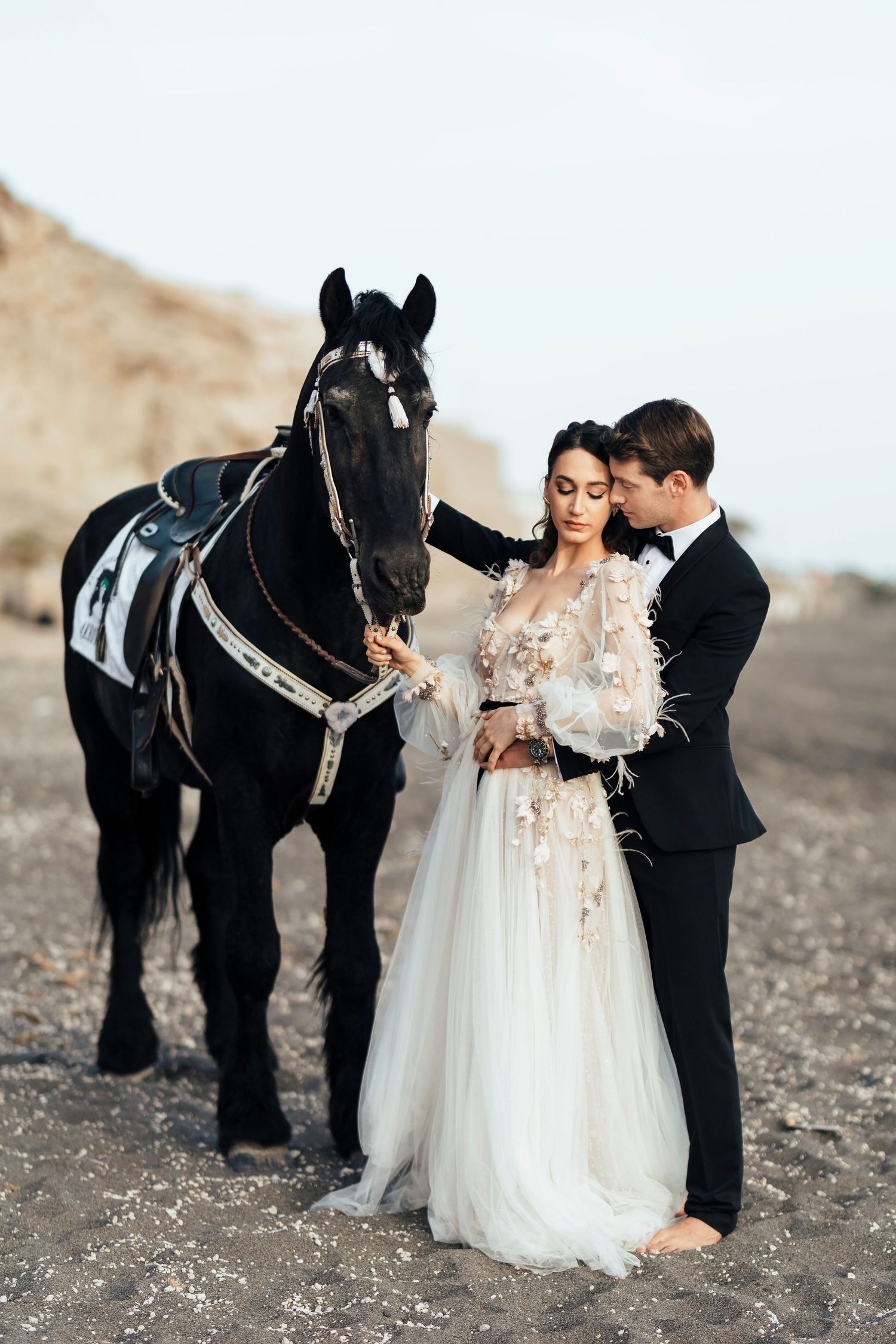 A couple in wedding attire stands beside a majestic black horse against a scenic desert landscape backdrop.