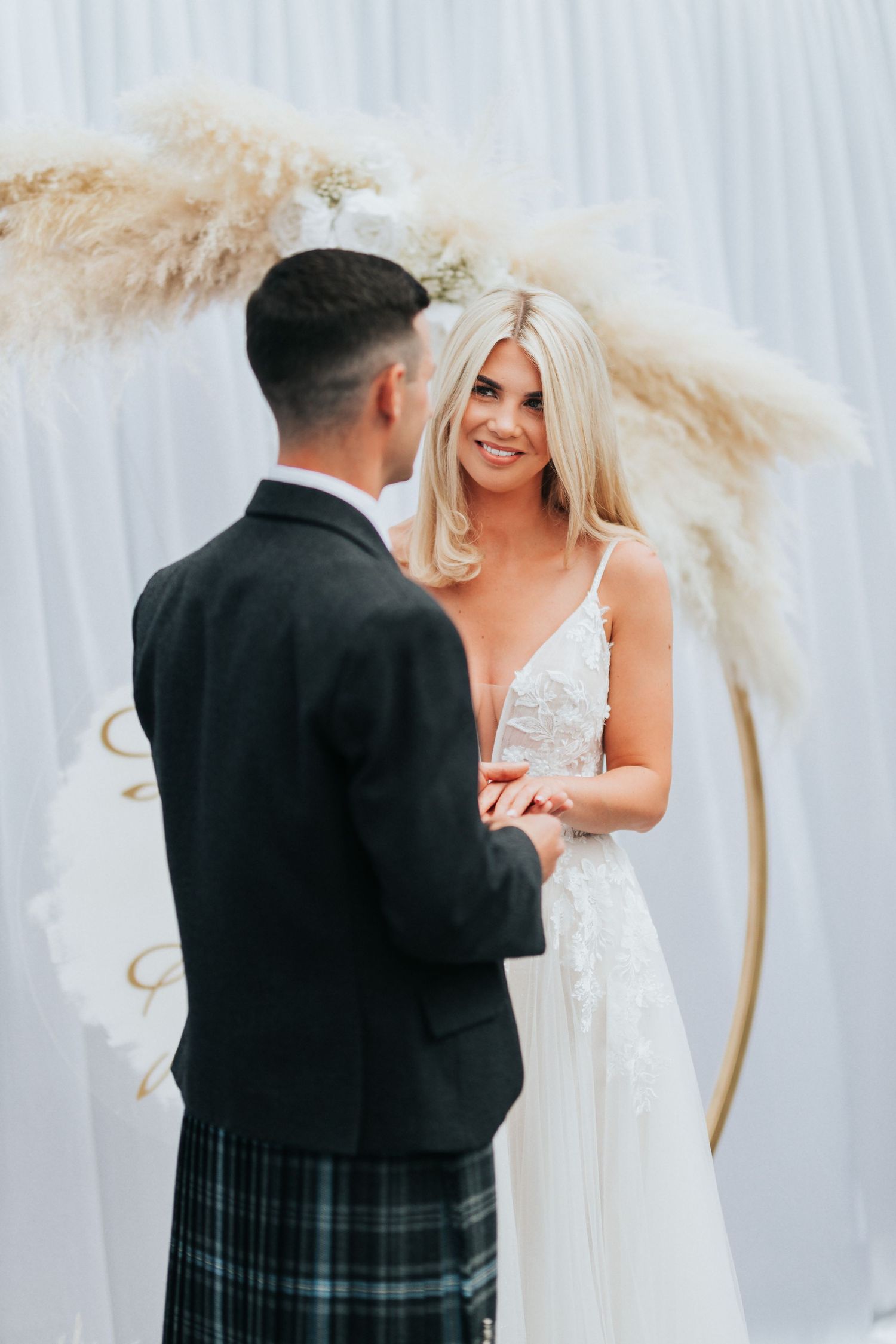 A wedding ceremony against a white backdrop decorated with pampas grass, featuring a couple exchanging vows.