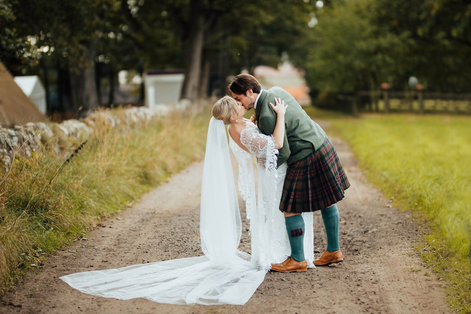 A couple in traditional Scottish wedding attire share a romantic moment on a country path.