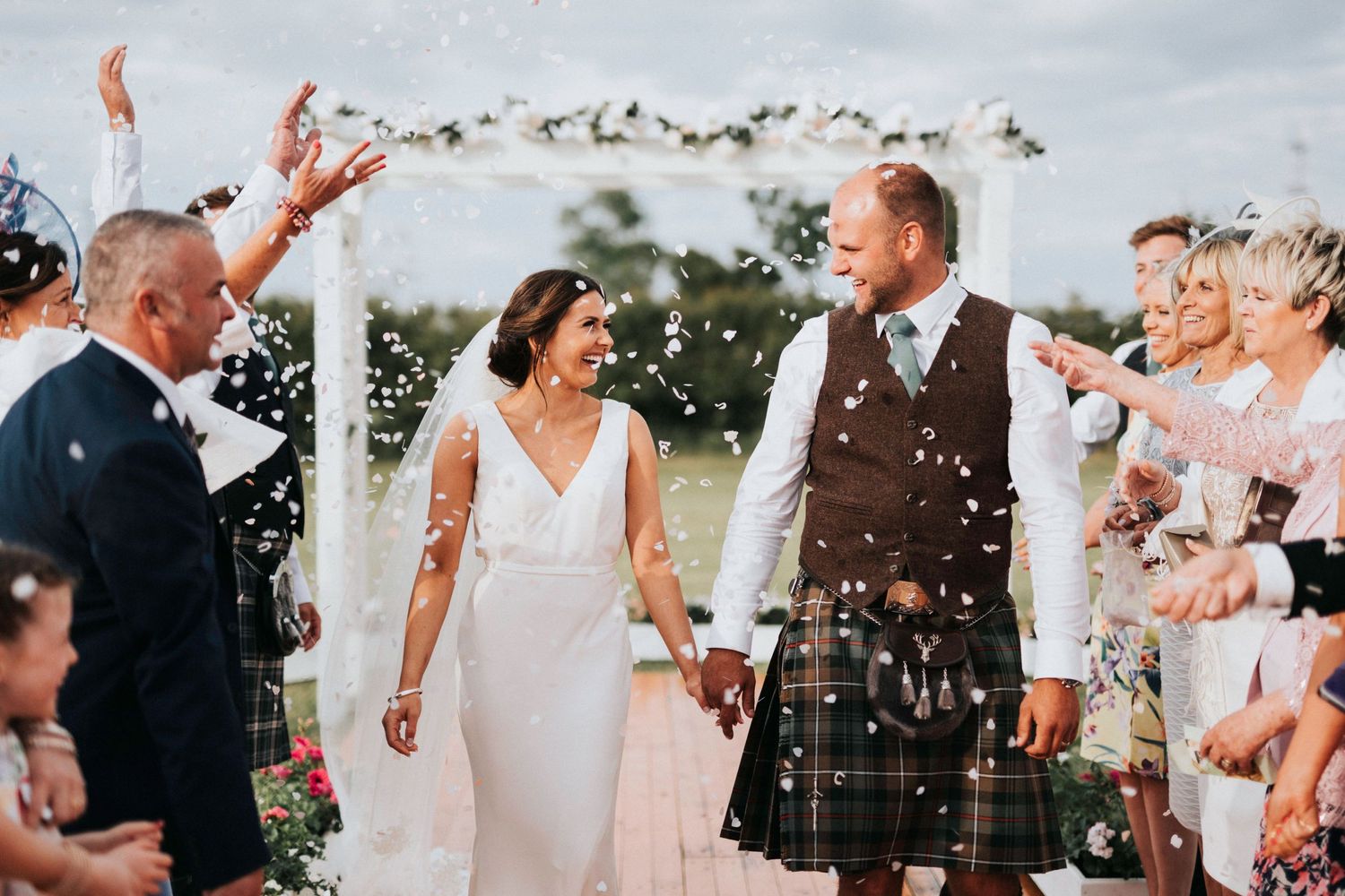 Wedding guests shower newlyweds with confetti during outdoor ceremony under floral arch at joyful celebration.