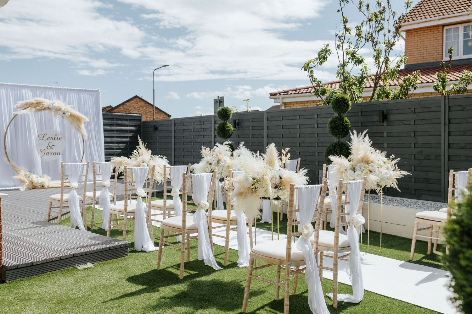 Elegant outdoor boho wedding ceremony setup with white curtains, dried pampas grass arrangements and garden backdrop.
