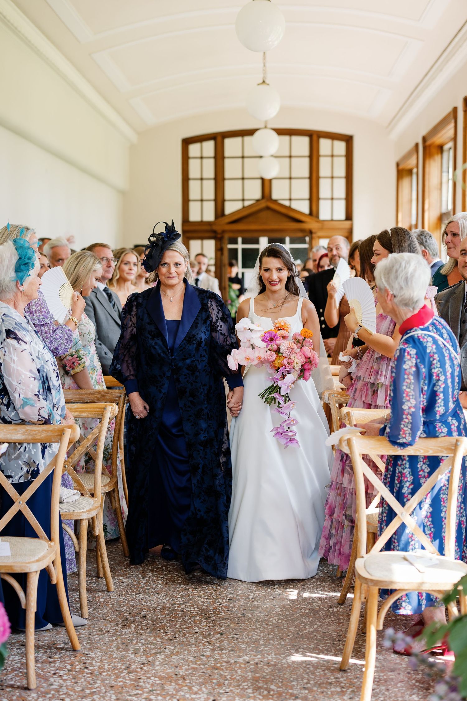 Wedding guests in elegant attire mingle and socialize at an indoor reception venue with wooden chairs and high ceilings.