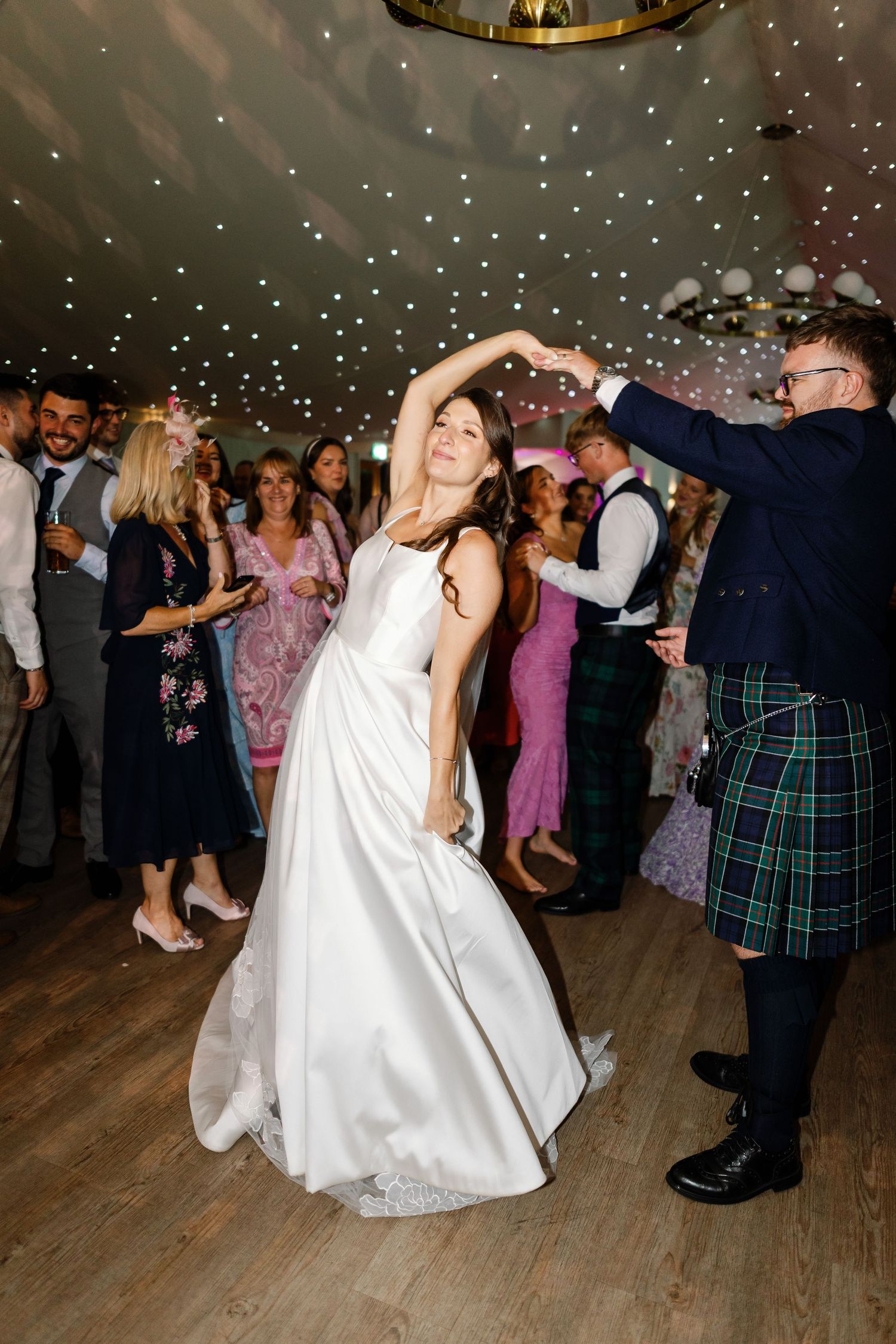 Wedding guests dance together on a wooden dance floor under twinkling lights during an evening reception celebration.