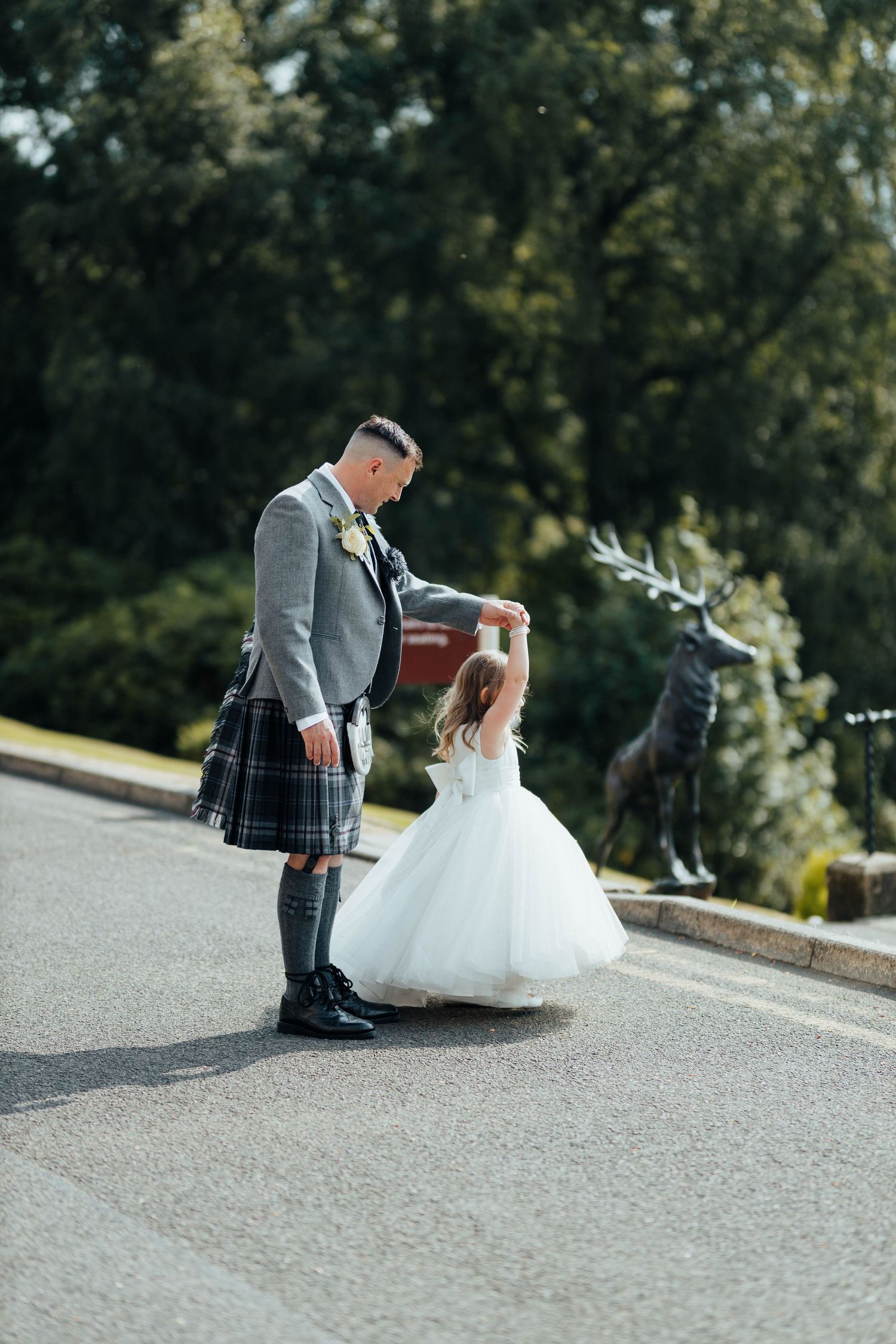 A person in traditional Scottish kilt and gray jacket twirls a young dancer in a white tulle dress on a paved road.