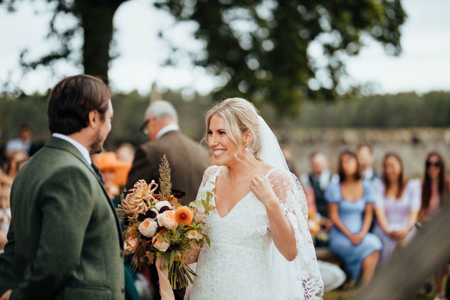 Outdoor wedding ceremony with bride in white lace dress holding fall-colored bouquet while guests watch in background.