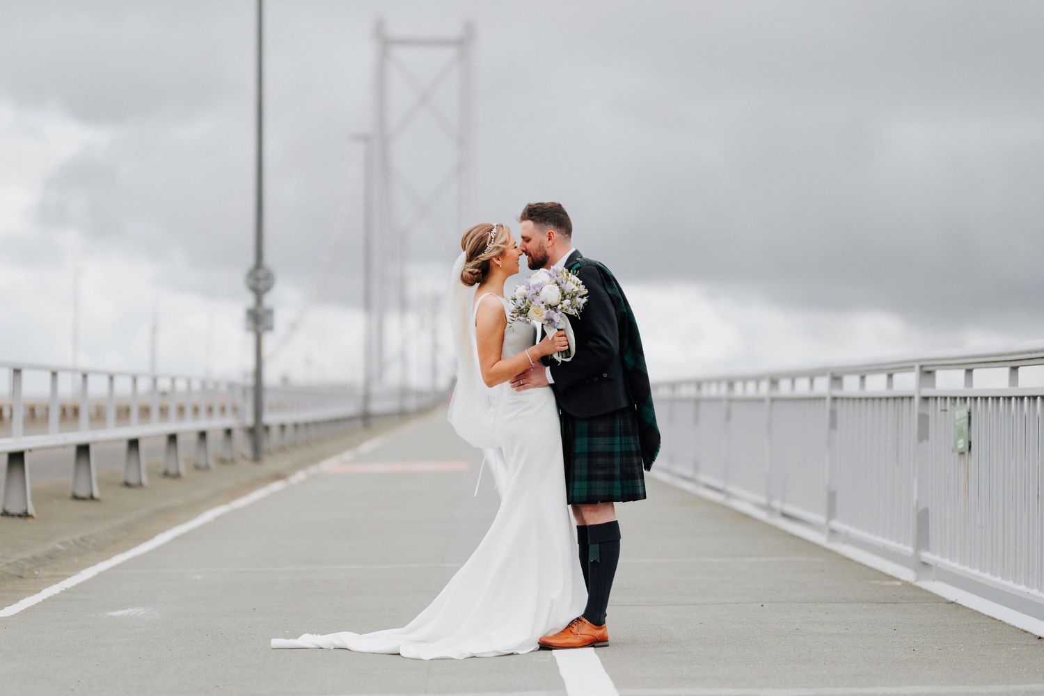 A couple shares a romantic moment on a foggy bridge walkway, with the bride in white and groom wearing a traditional kilt.